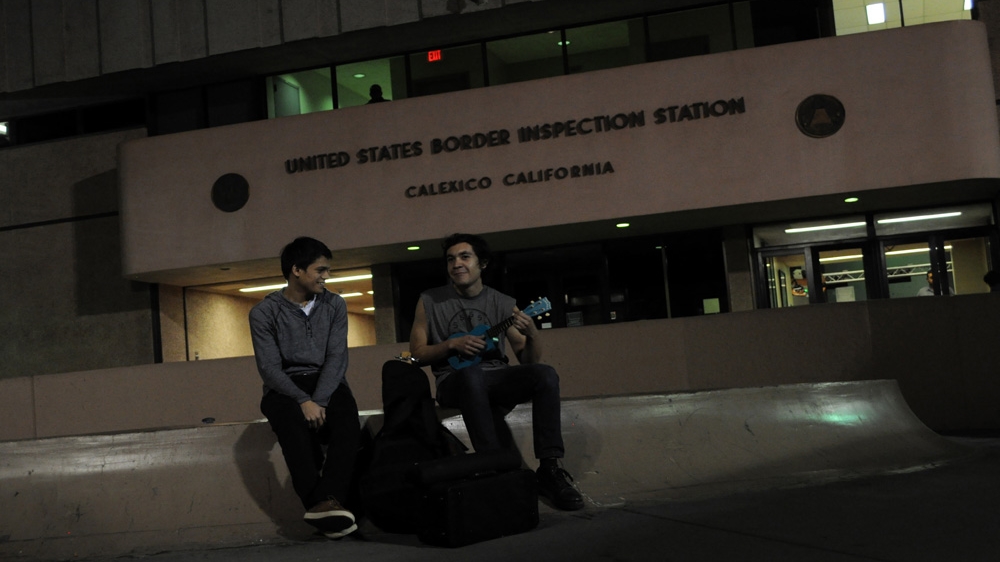 A man plays guitar, seated next to a friend, outside the US-Mexico pedestrian border crossing in Calexico, California [Joe Jackson /Al Jazeera] 