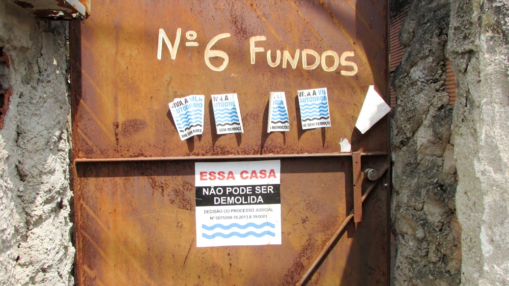 The remaining houses in the community bear these stickers. The top ones are the Vila Autodromo resistance movement's logo. They read 'Long live Vila Autodromo! Rio without Removals'. The sticker below says 'This house must not be demolished,' and cites a judicial ruling [Maya Thomas-Davis/Al Jazeera]