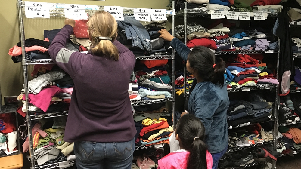 A volunteer helps a mother and daughter to pick out clothing from a rack of donations, organised by size and type, at Annunciation House [Sophie Eastaugh/Al Jazeera] 