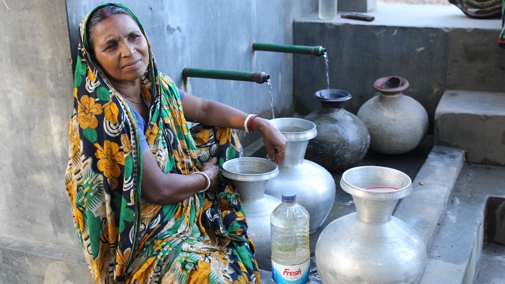 Savitri Mundo waits by the water pump in Fultala. Her husband, Bibhuti, now works in Dhaka as a security guard and comes home every three months [Neha Thirani Bagri/The GroundTruth Project]