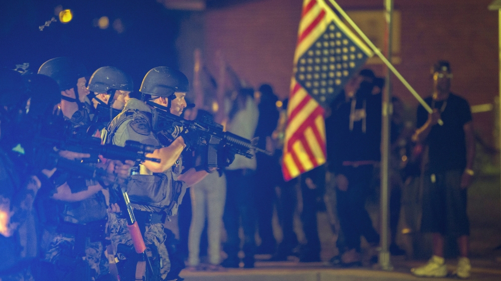 A police officer raises his weapon at a car speeding in his general direction as a more vocal and confrontational group of demonstrators stands on the sidewalk during further protests in reaction to t