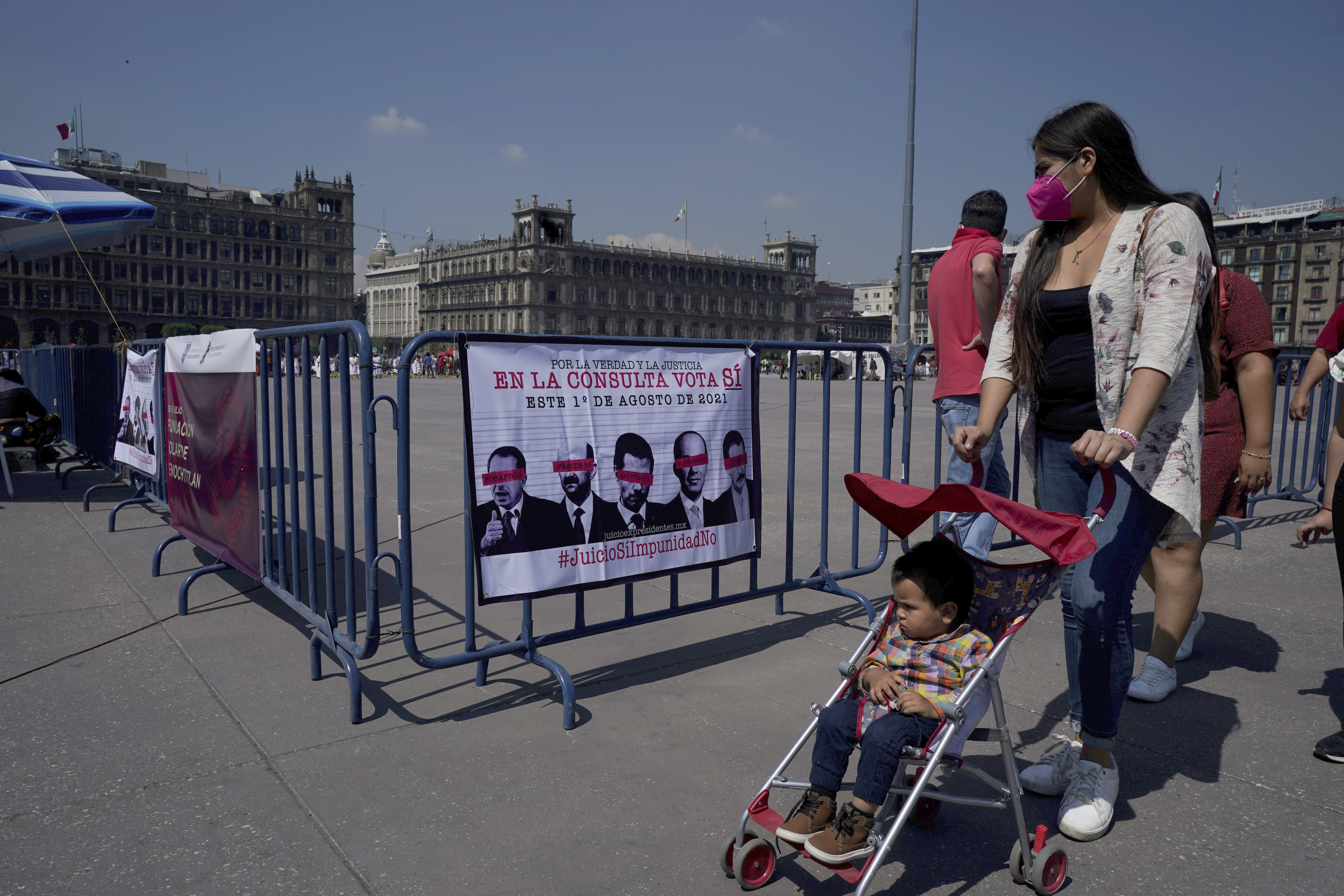 In Mexico City's main square, the Zocalo, a woman pushes a stroller past a sign showing images of several Mexican former presidents and calling for citizens to participate in a referendum on whether ex-presidents should be tried for their alleged crimes during their time in office [File: Fernando Llano/AP Photo]
