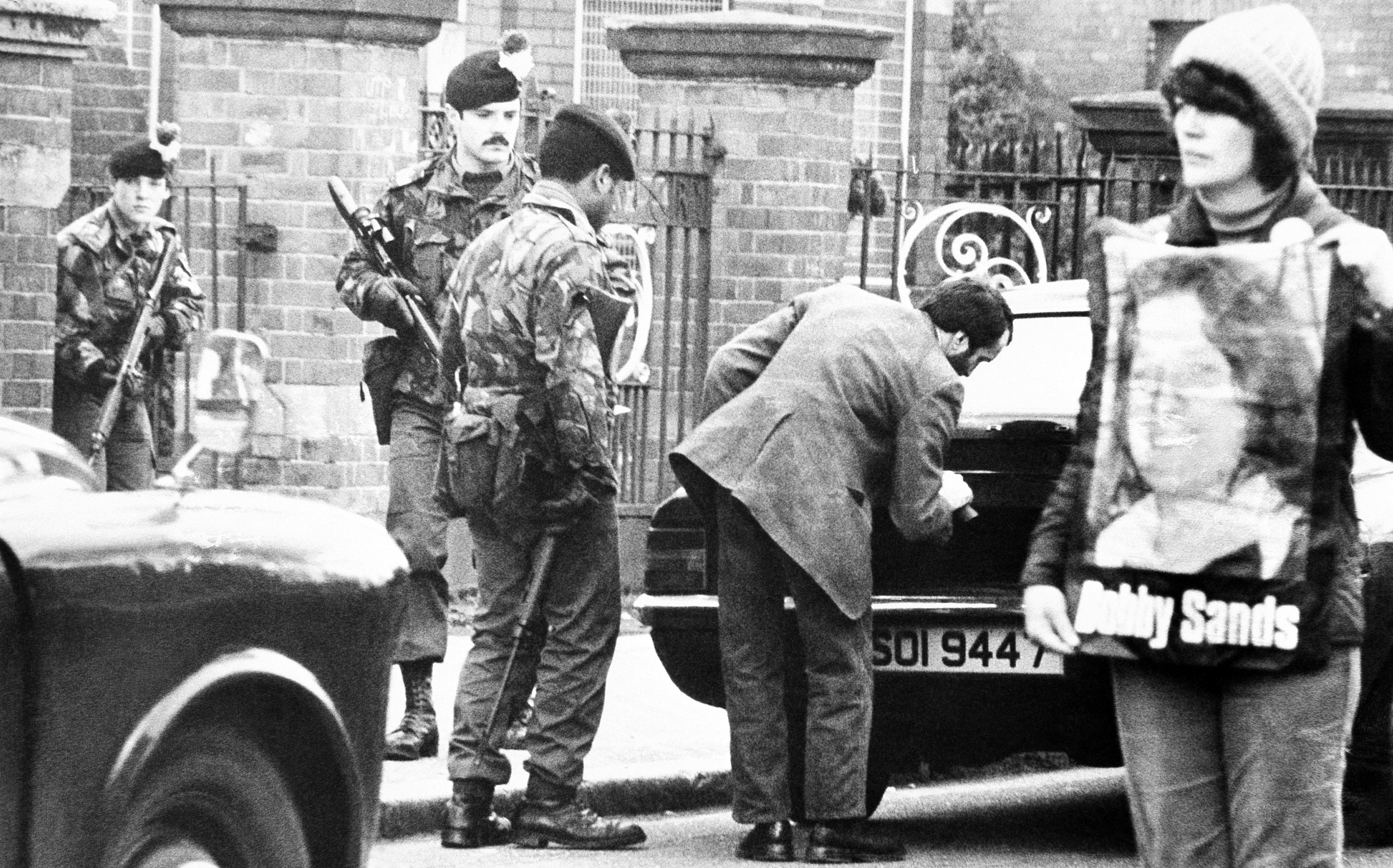 A protester supporting the IRA hunger strikers stands in the centre of the Falls Road in Belfast on May 1, 1981, as British soldiers check cars at a roadblock [File: AP Photo/Peter Kemp]