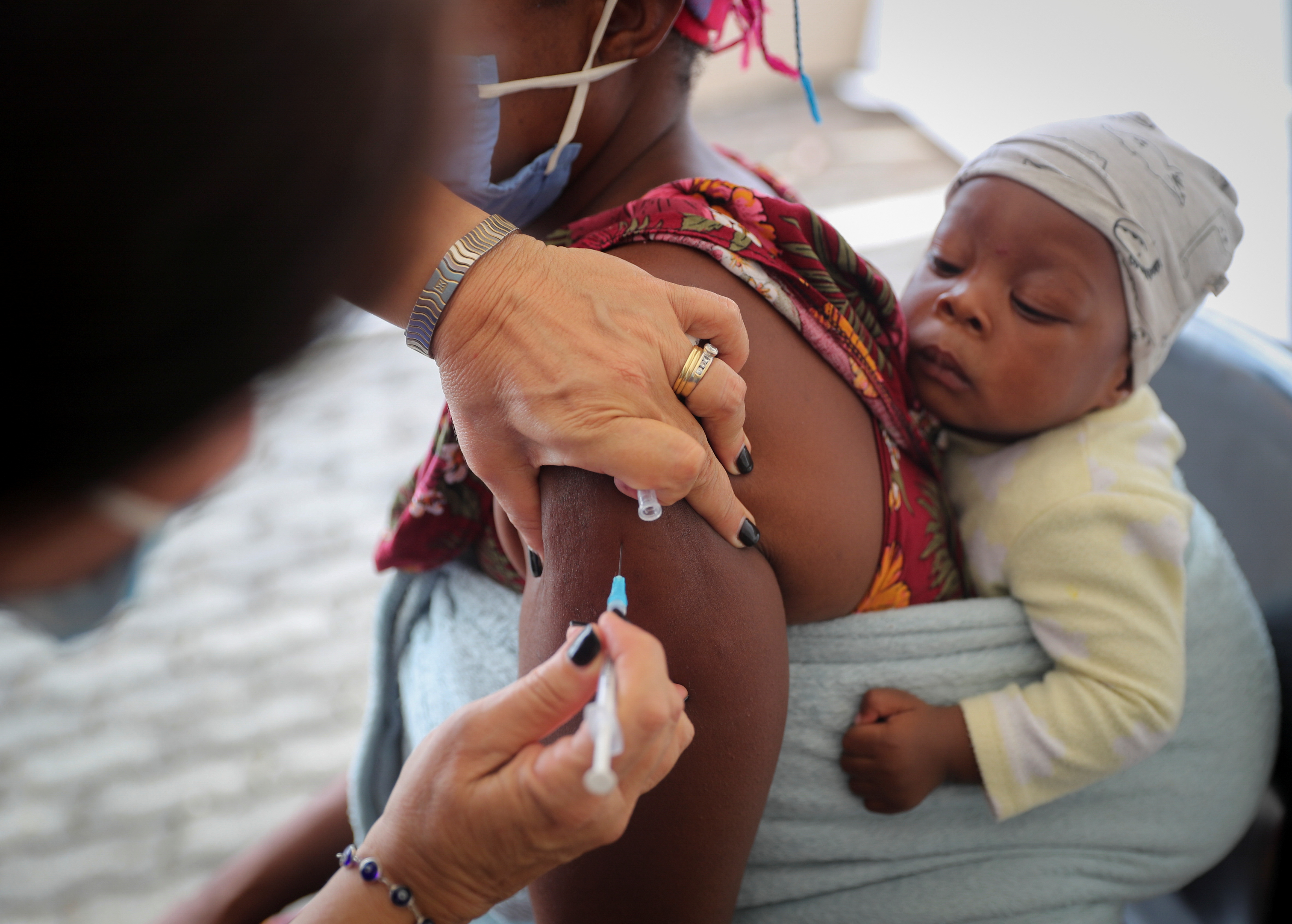 A healthcare worker administers a dose of COVID-19 vaccine to a woman, as her baby tied to her back watches on.