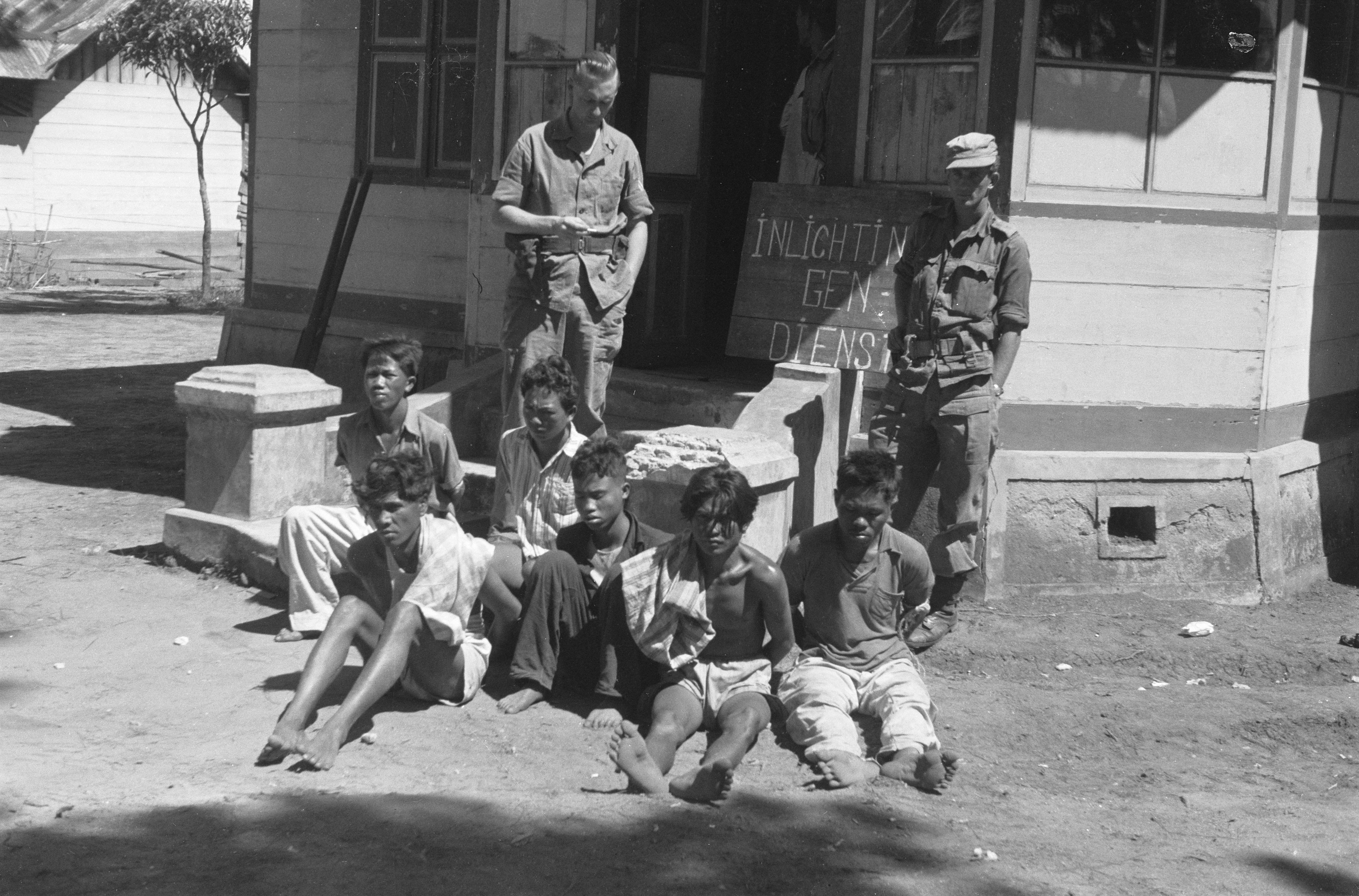 Two Dutch soldiers stand on the veranda of a house watching over a group of six Indonesian independence fighters who they plan to interrogate in 1946
