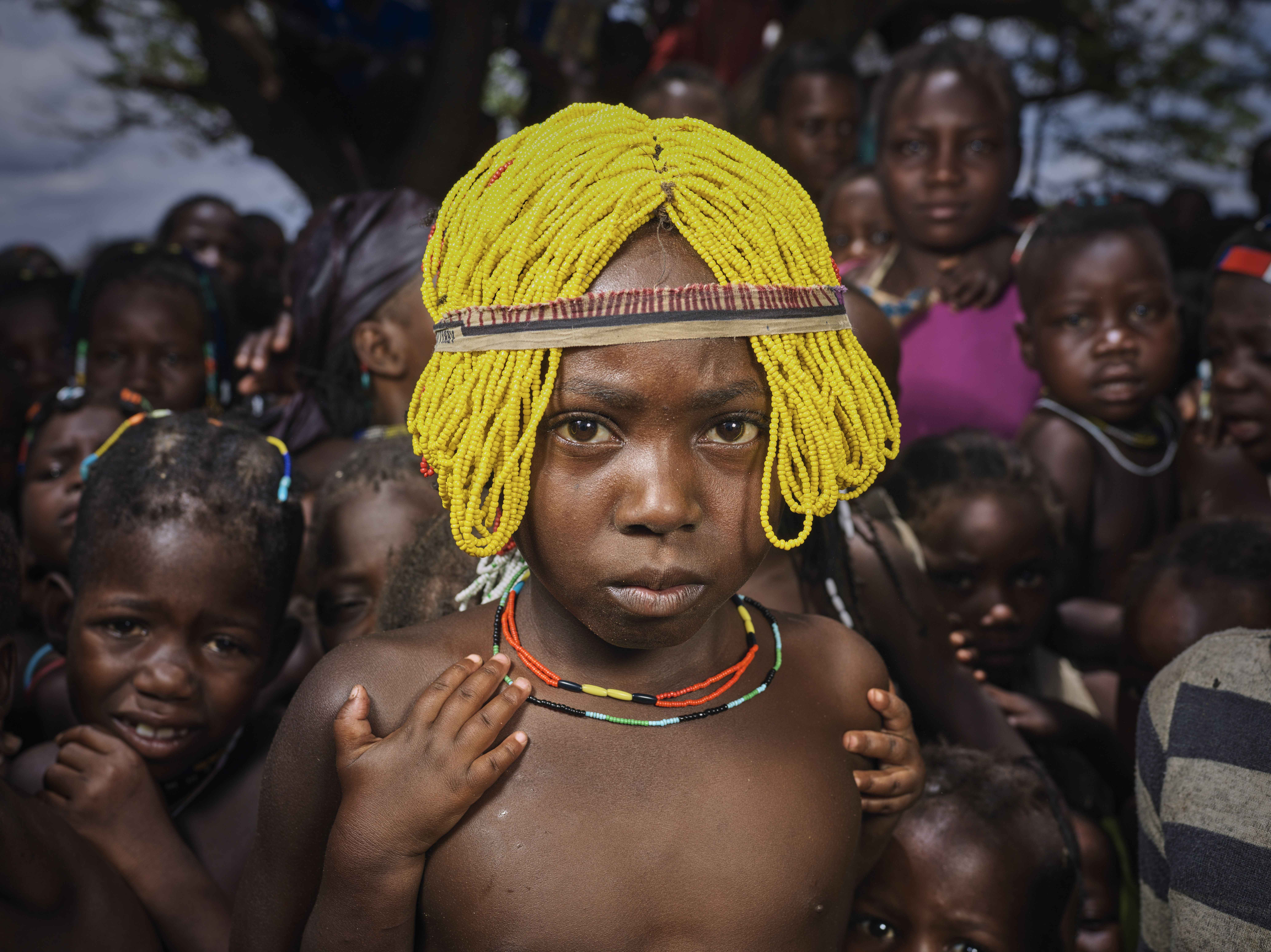 A photo of a refugee girl wearing a traditional beaded wigs called Ena meant for girls of the Mudimba tribe from southern Angola with a group d children in the back.