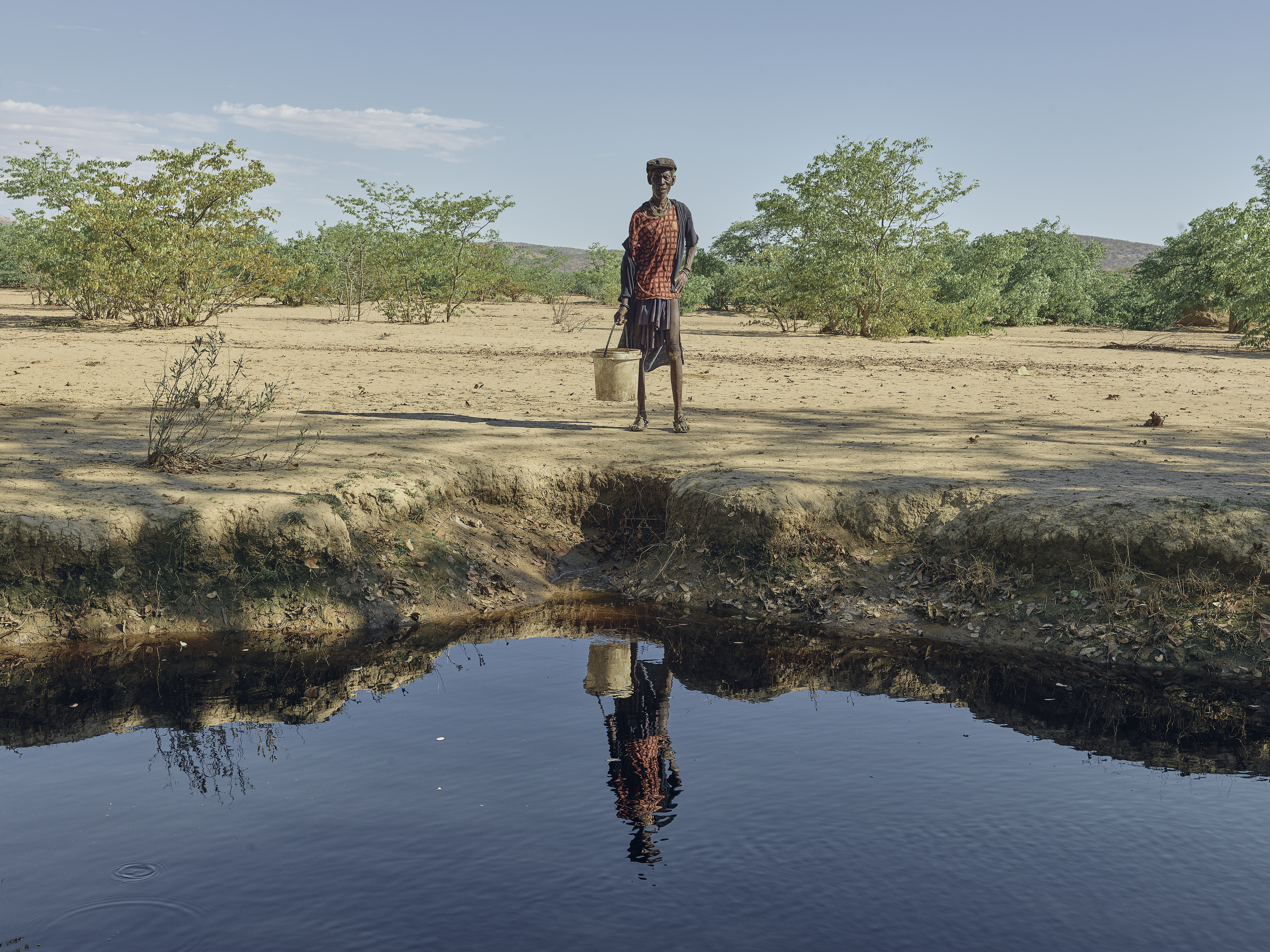 A photo of a man standing in front of a river looking at the camera on the other side of the river.