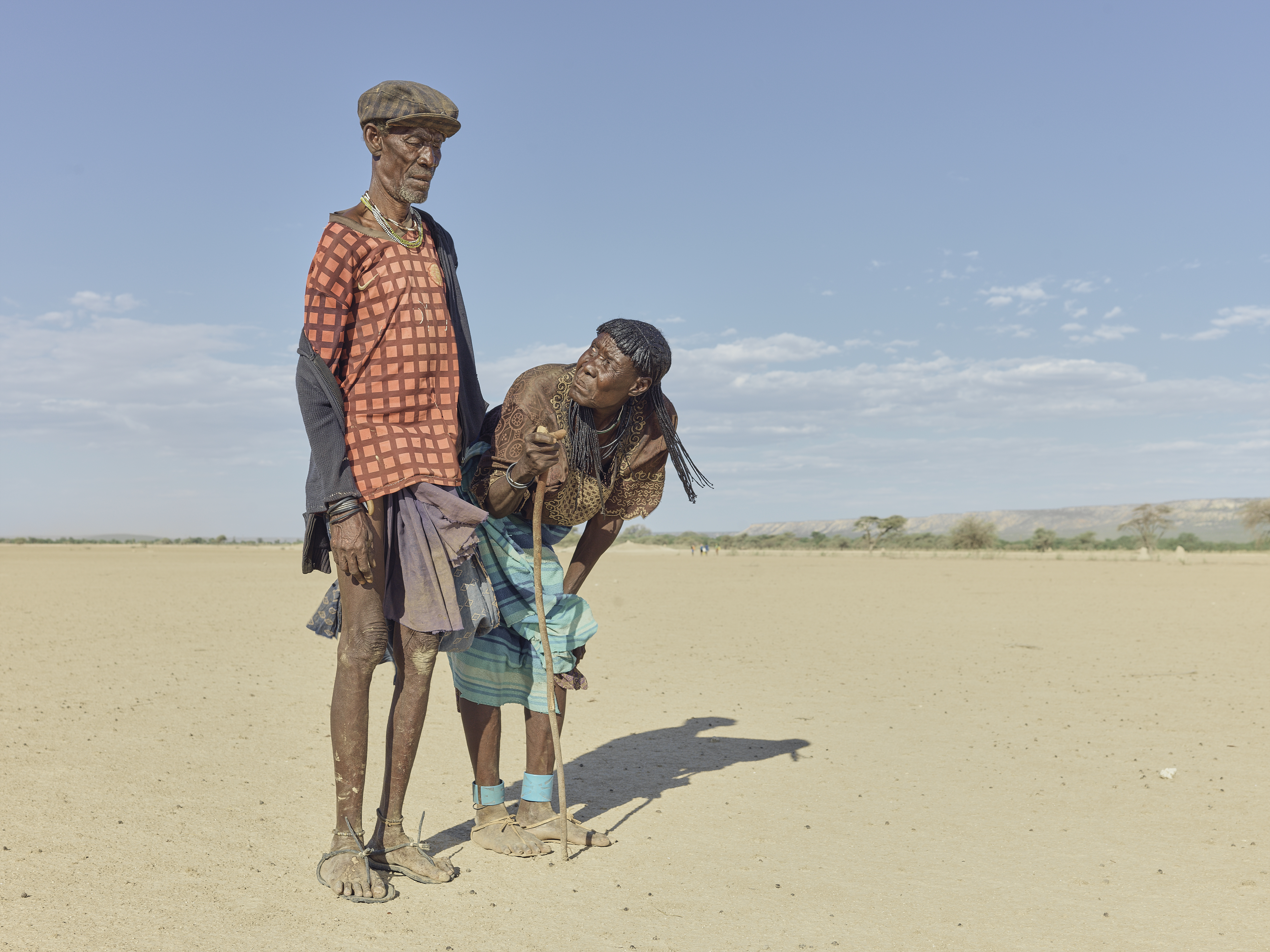 A photo of two people next to each other, side by side , one of them is holding a cane and bending down looking up at the other from the side.