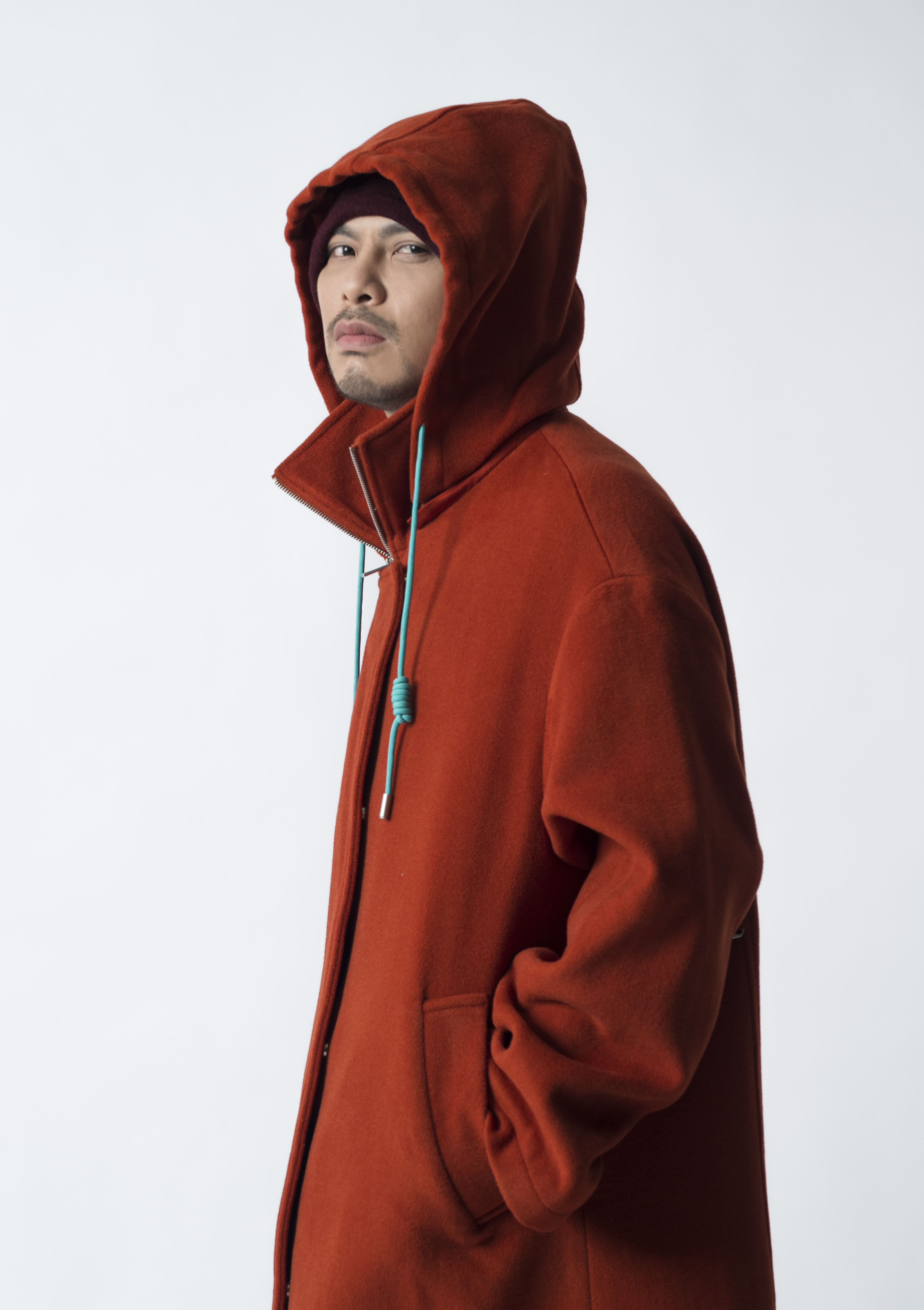 A publicity photo of Malaysian performer Namewee dressed in a red hoodie with the hood pulled up and his hands in his pockets