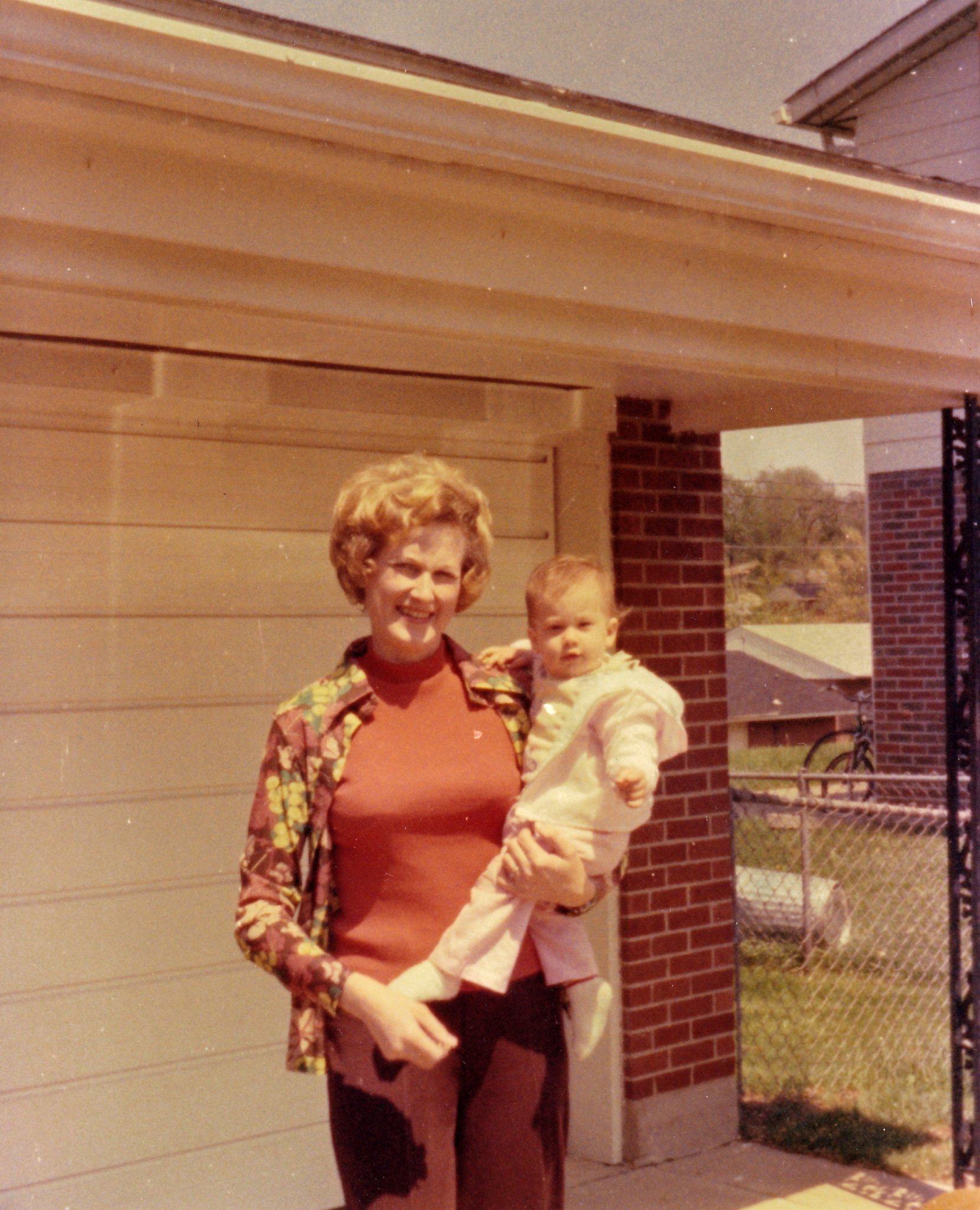 The writer's mother carrying her as a baby, standing in front of a house on a bright sunny day.