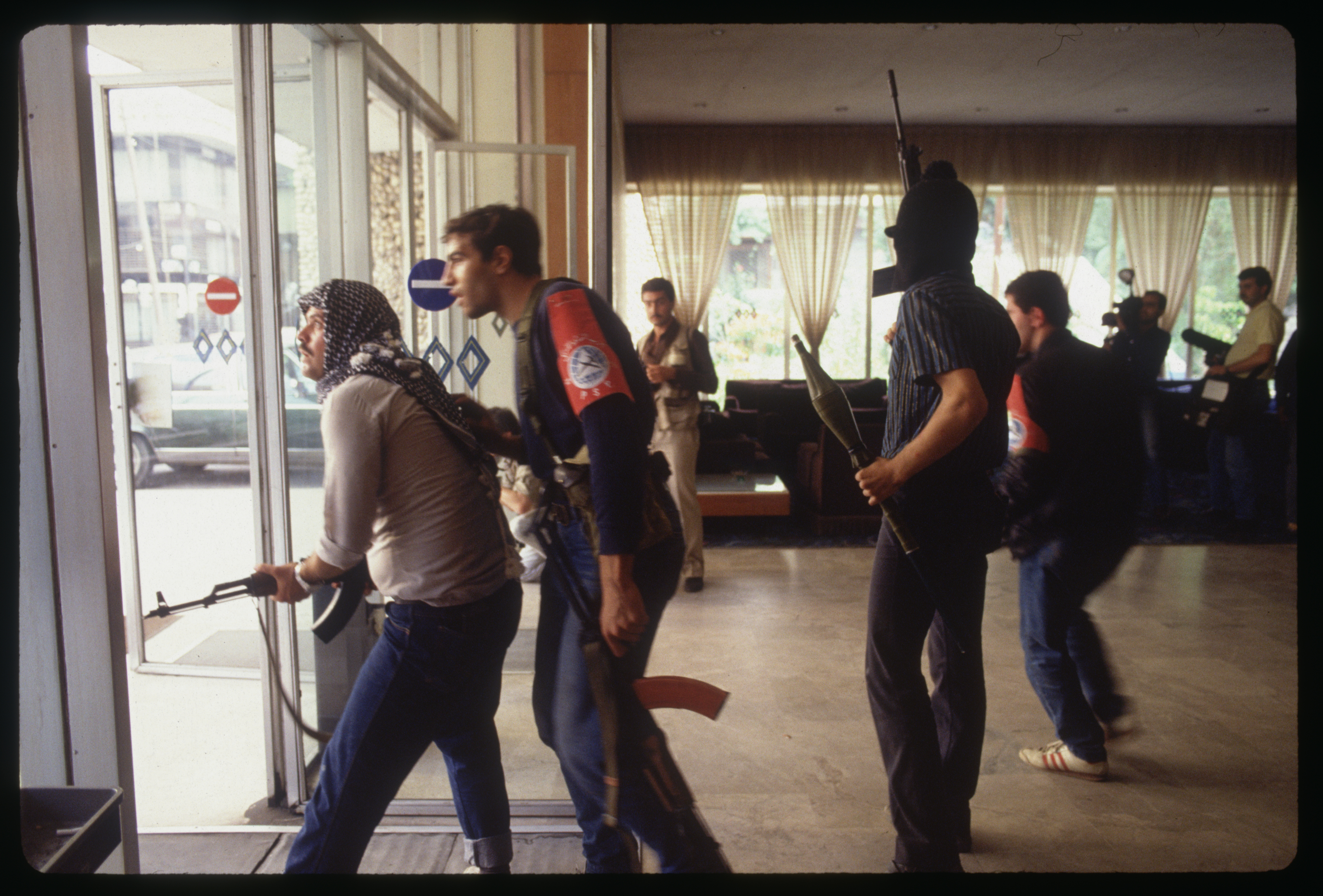 Several armed men stand in the lobby of a hotel looking out through the glass doors and windows to the street beyond