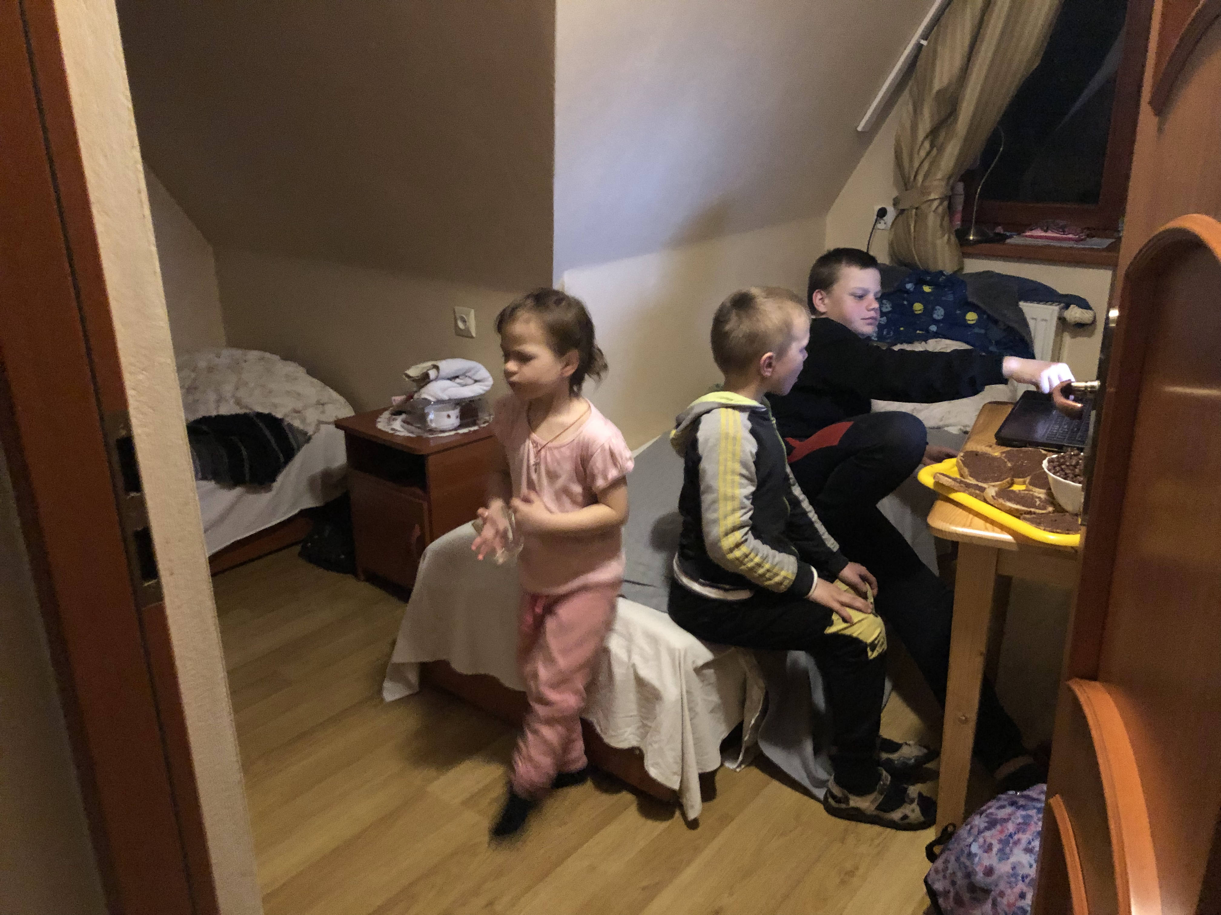 A photo of three children sitting in a room. one is on the computer, the other is sitting next to him and the third child is standing behind them.