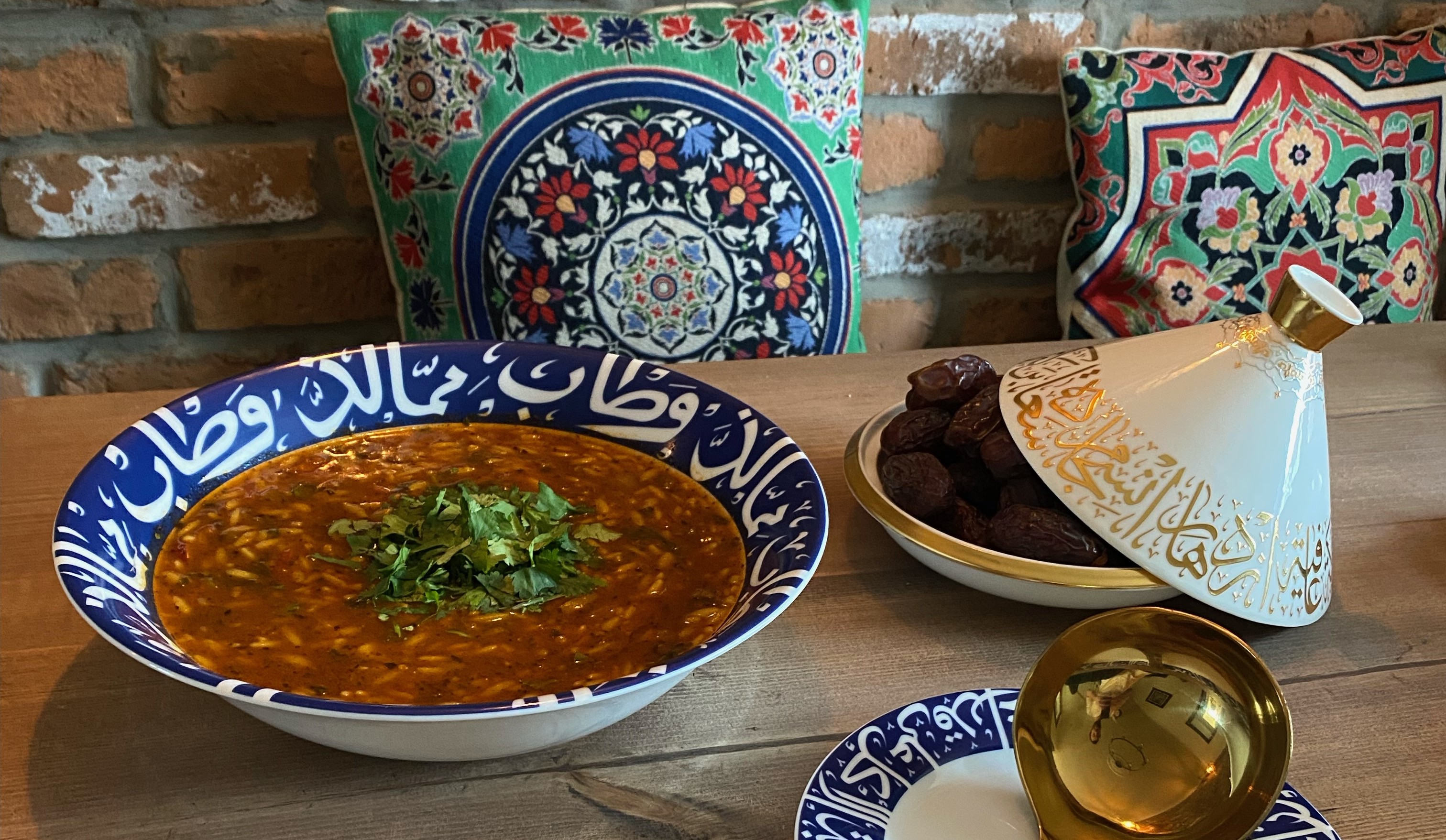 A bowl of sherba and a bowl of dates with a golden-coloured ladle on a wooden table