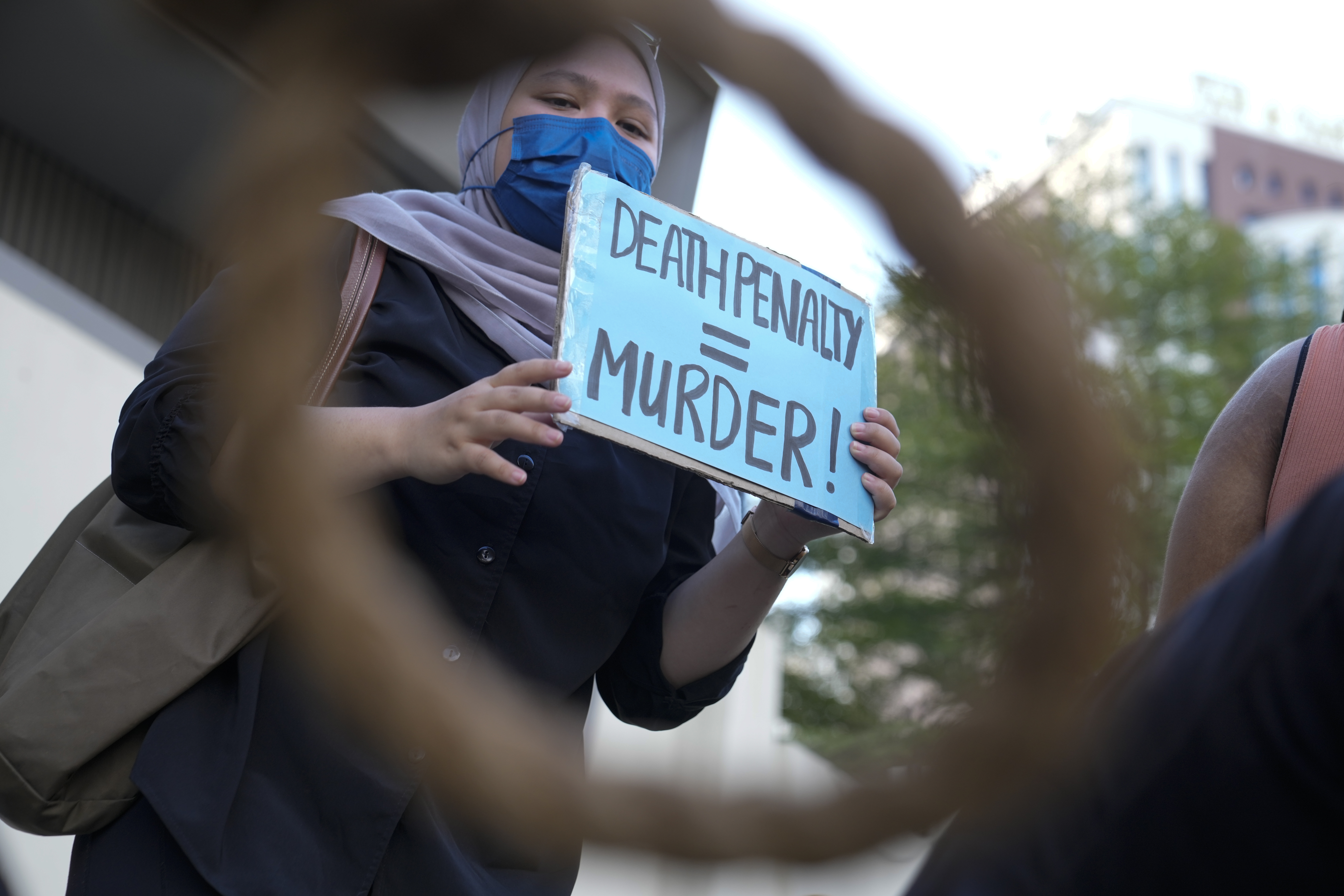 A young woman, seen through a blurry noose, holds up a banner saying 'death penalty = murder'.