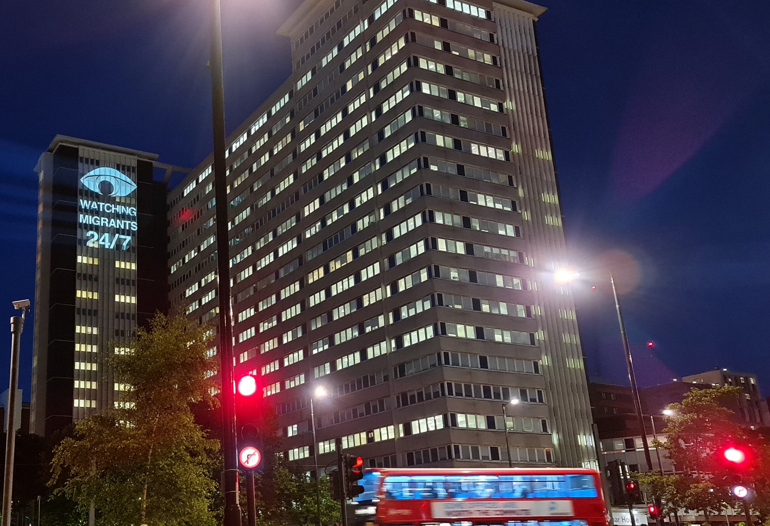 A light projection on a Home Office Immigration reporting centre reads 'watching migrants 24/7'
