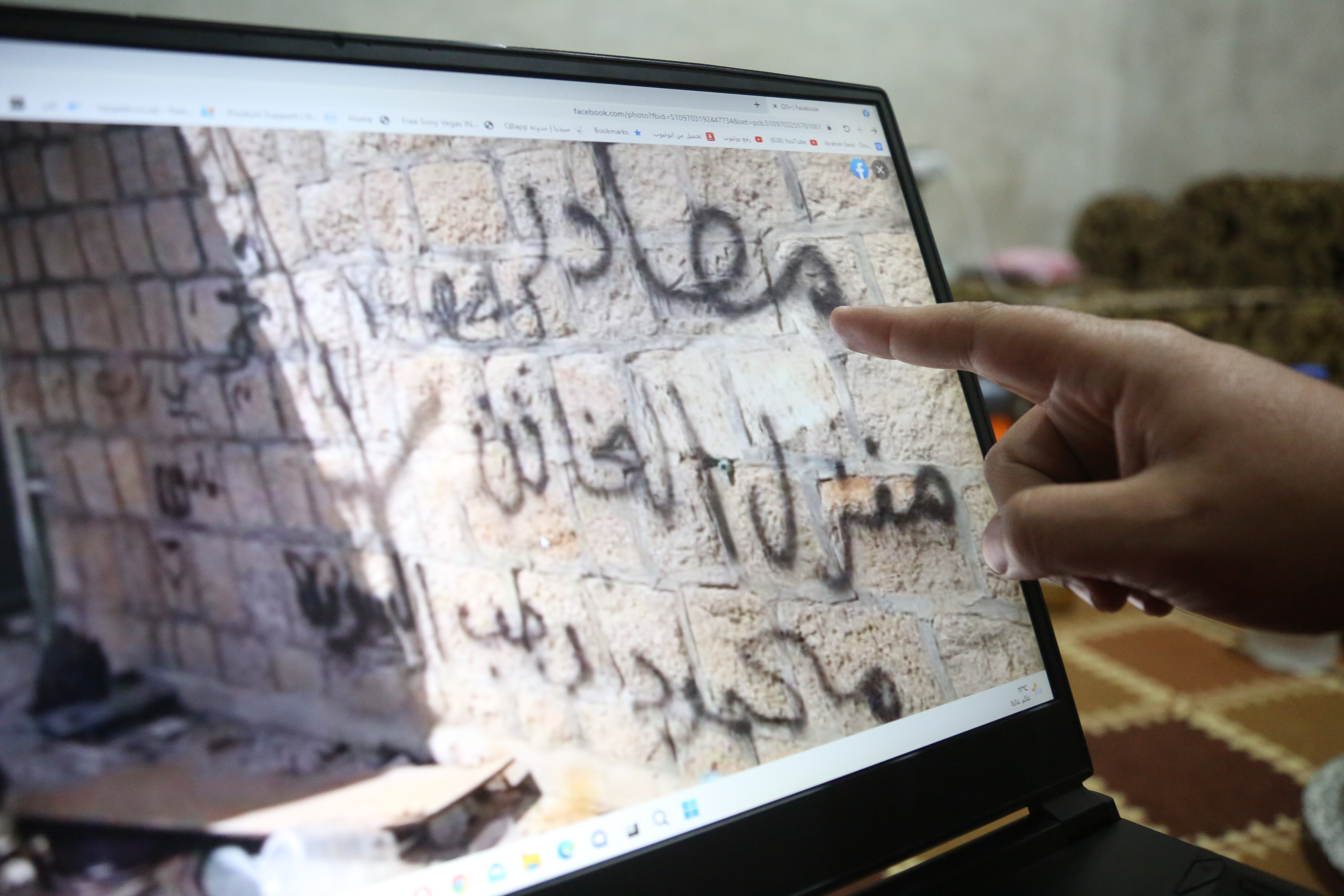 A man points to a screen showing a picture of a wall. Graffiti on the wall says 'Confiscated home of a traitor'
