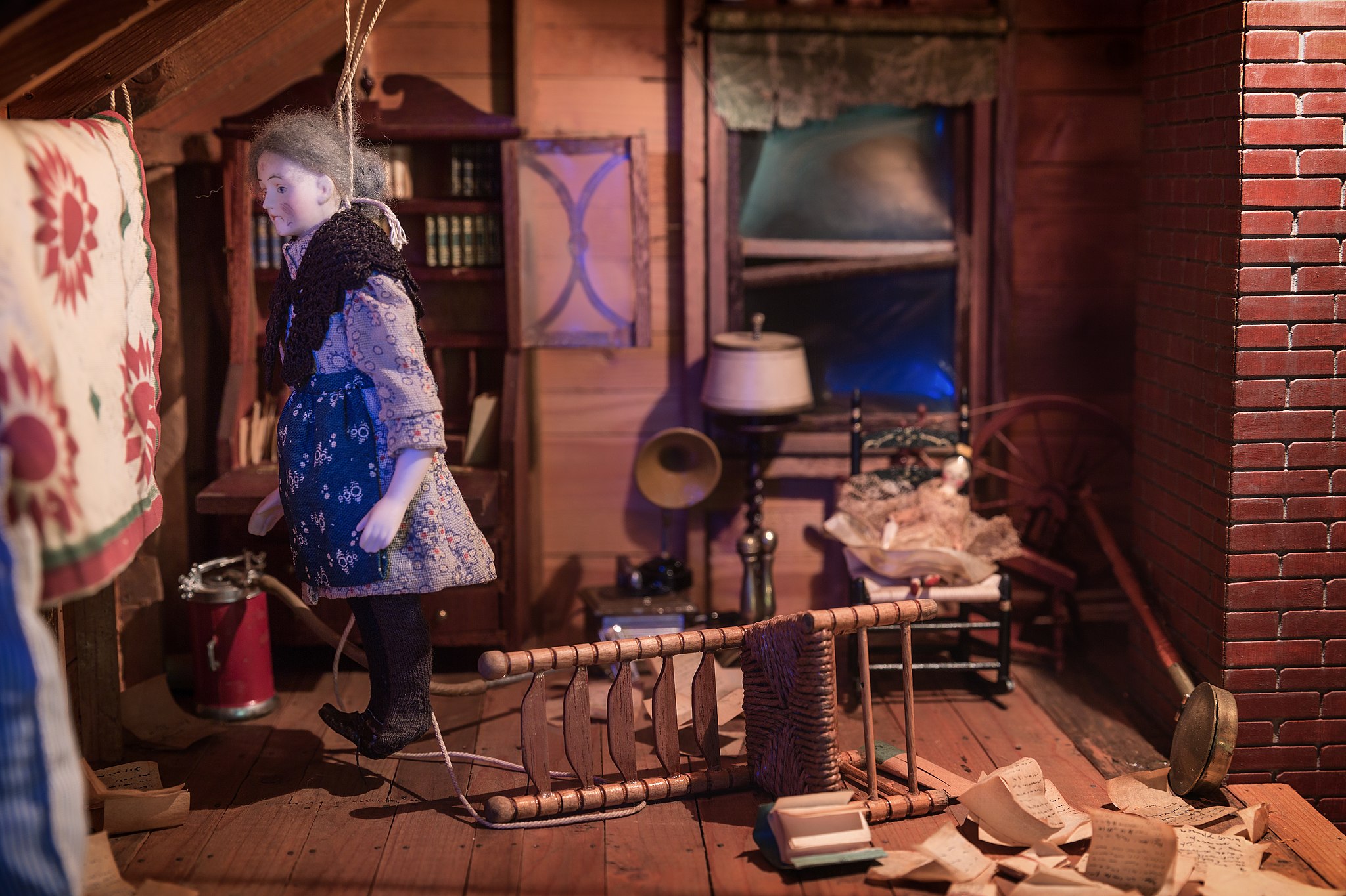 A photo of a diorama of a doll standing in front of a mirror with a chair on the ground behind it and papers on the floor.