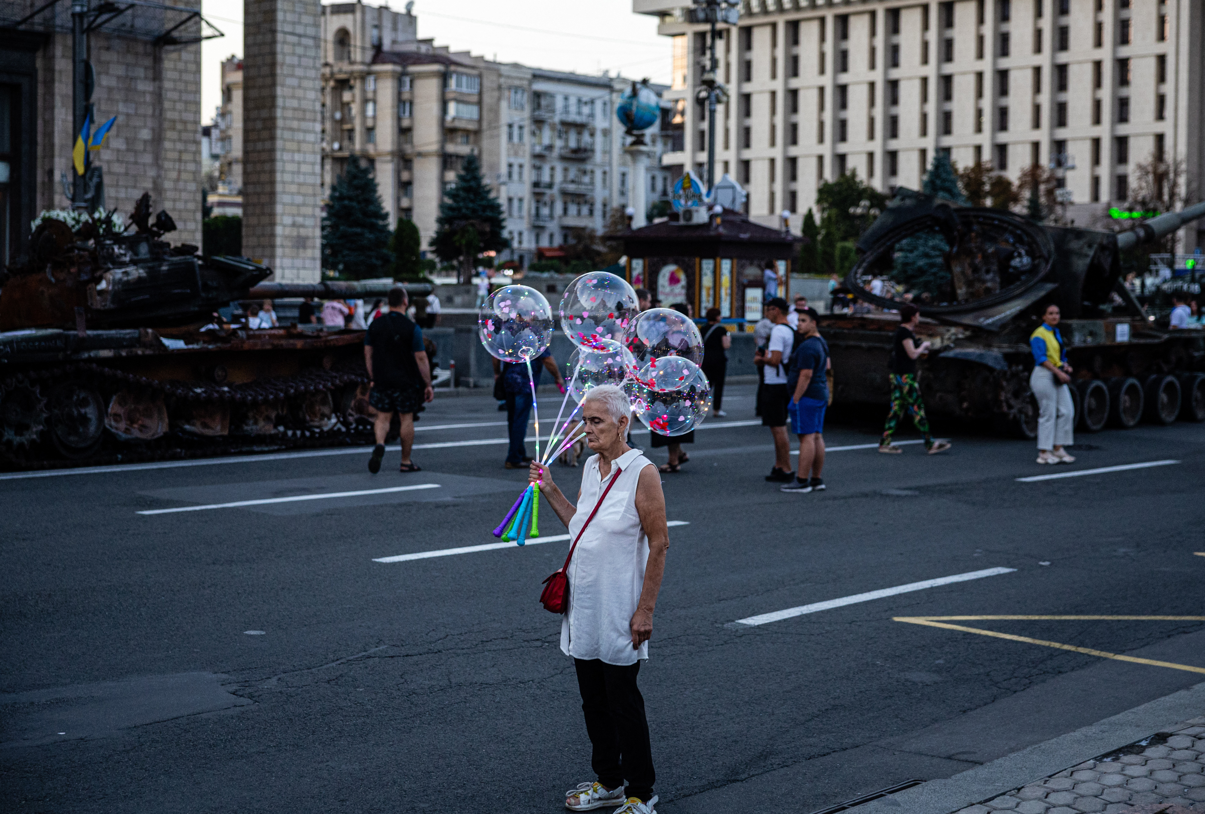 An elderly woman sells glowing balloons next to a destroyed Russian military equipment in Kyiv, ahead of Ukraine's Independence Day.