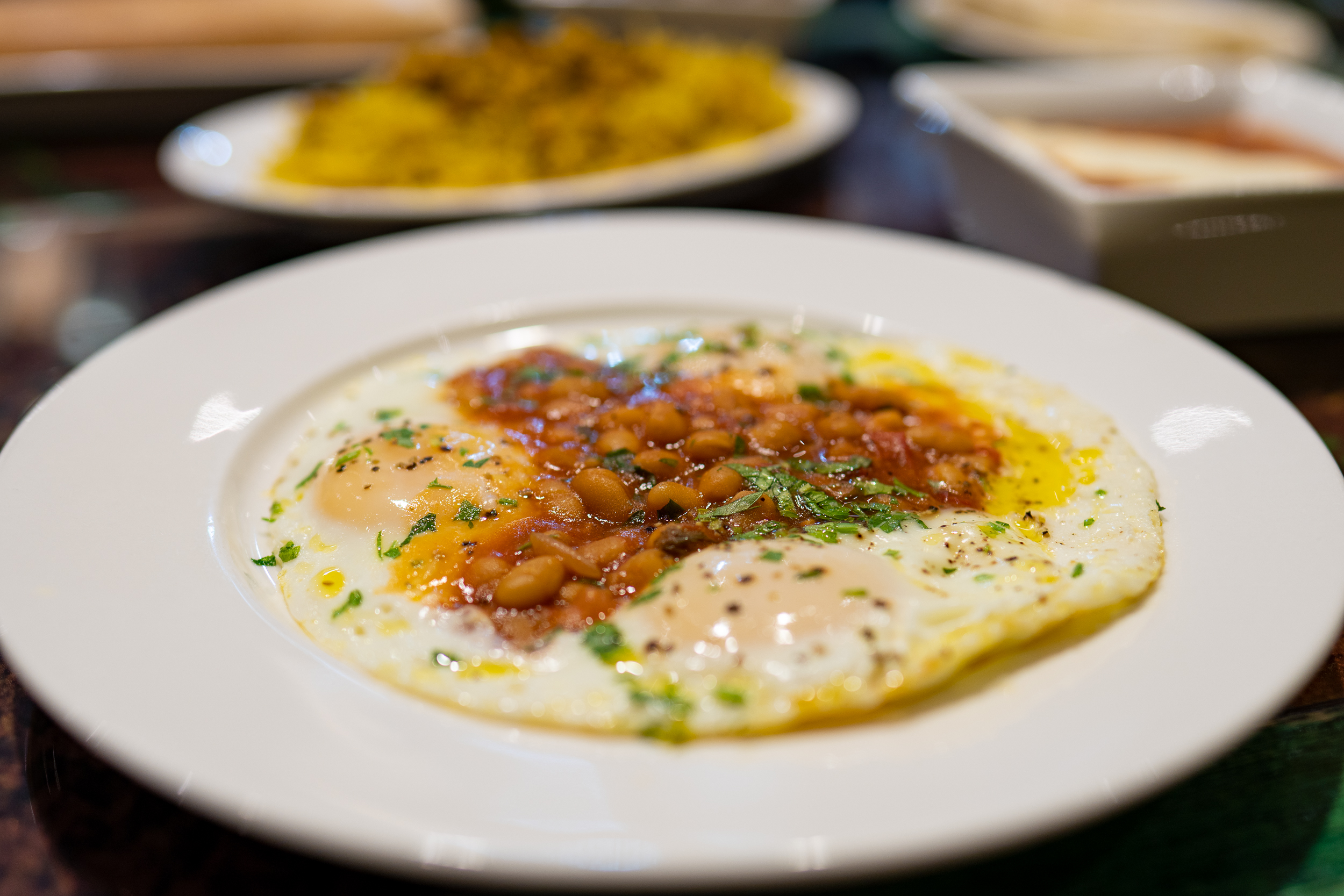 Three sunny-side up eggs on a shite plate with a dollop of white beans in a tomato sauce on top, and chopped herbs sprinkled over top