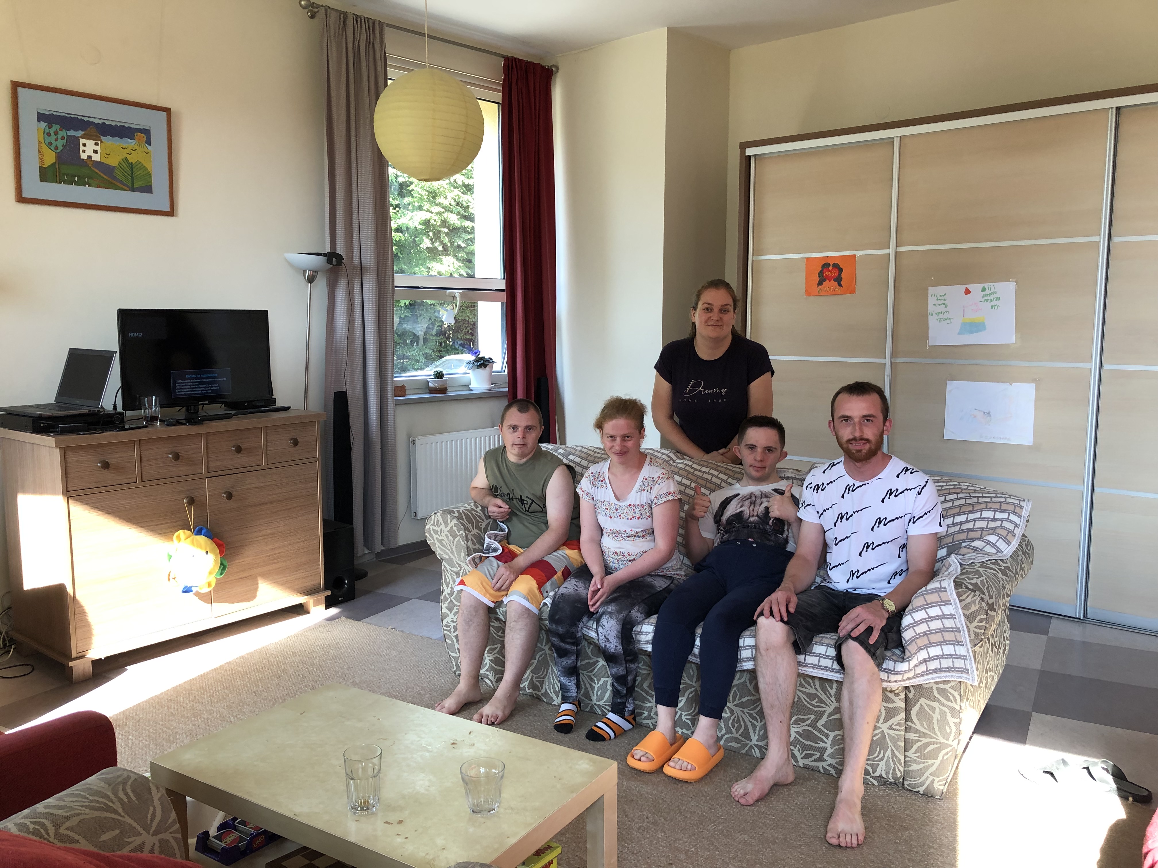 A photo of 5 people, 4 sitting on a sofa and one standing behind them, in a room with a television and a coffee table in front of them.