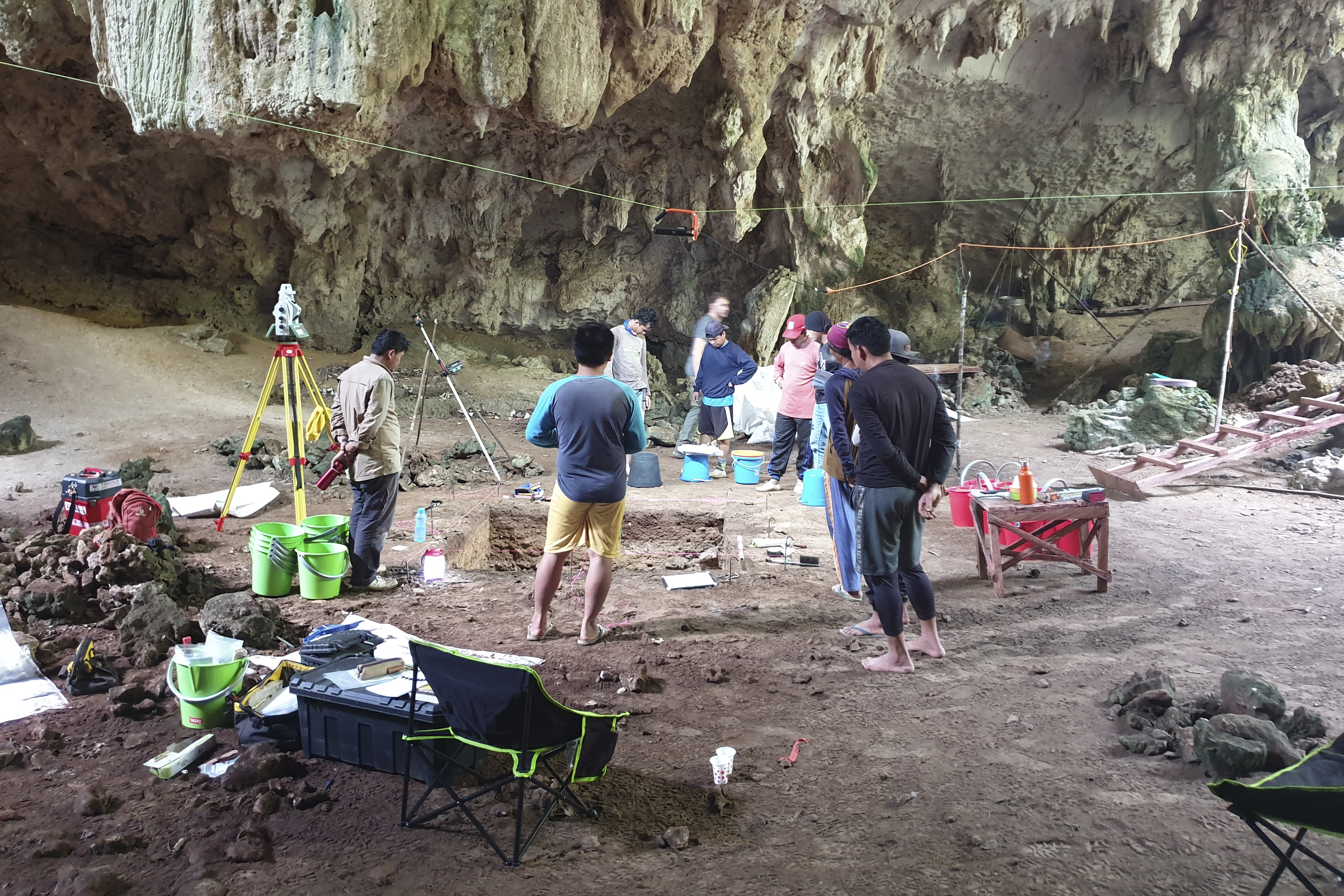 Researchers inside the Liang Tebo cave gathered around the pit with the skeleton inside, with camping chairs, buckets and other equipment on the cave floor around them. The interior walls of the cave are visible behind them
