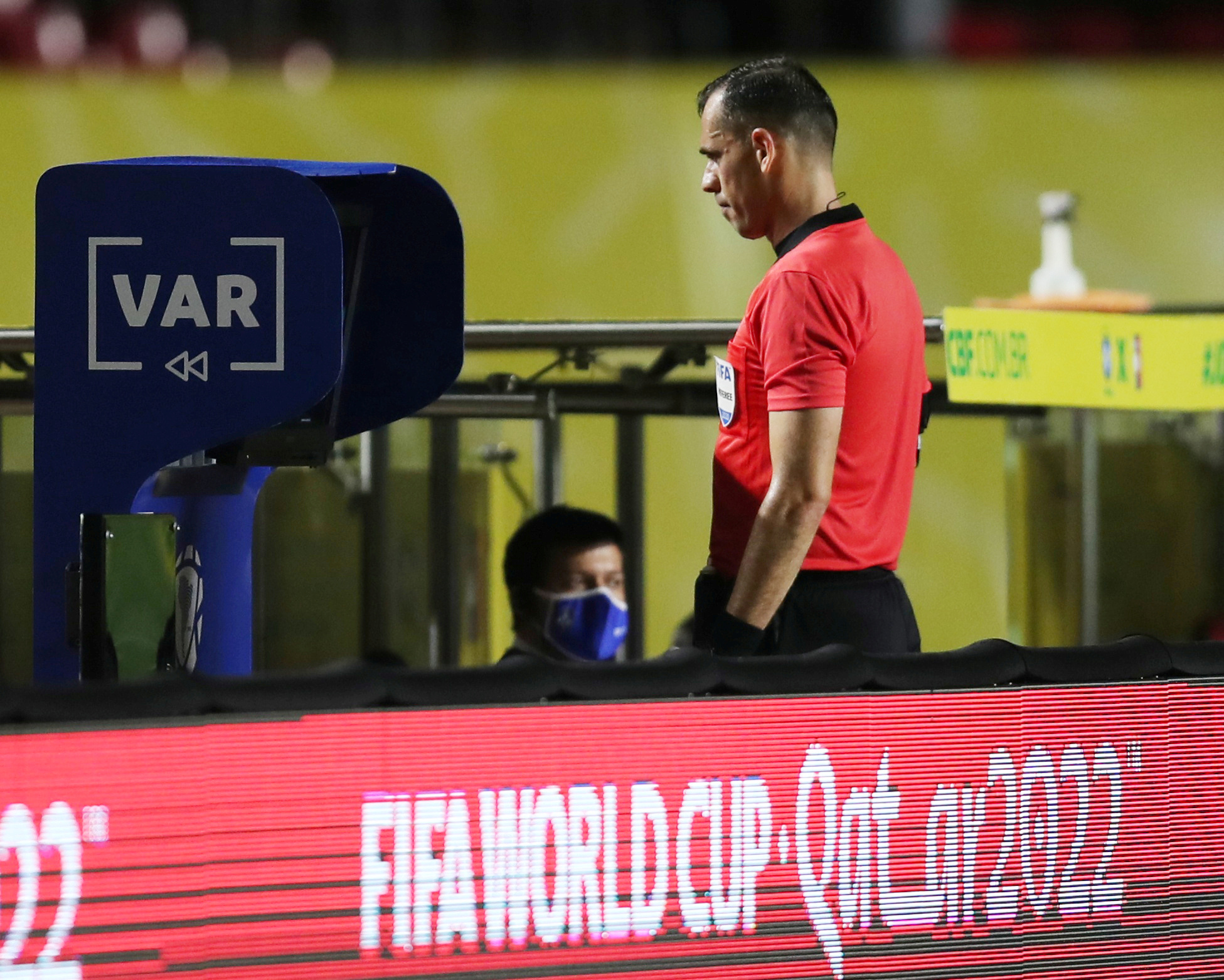 A referee in red t-shirt stands in front of a video screen
