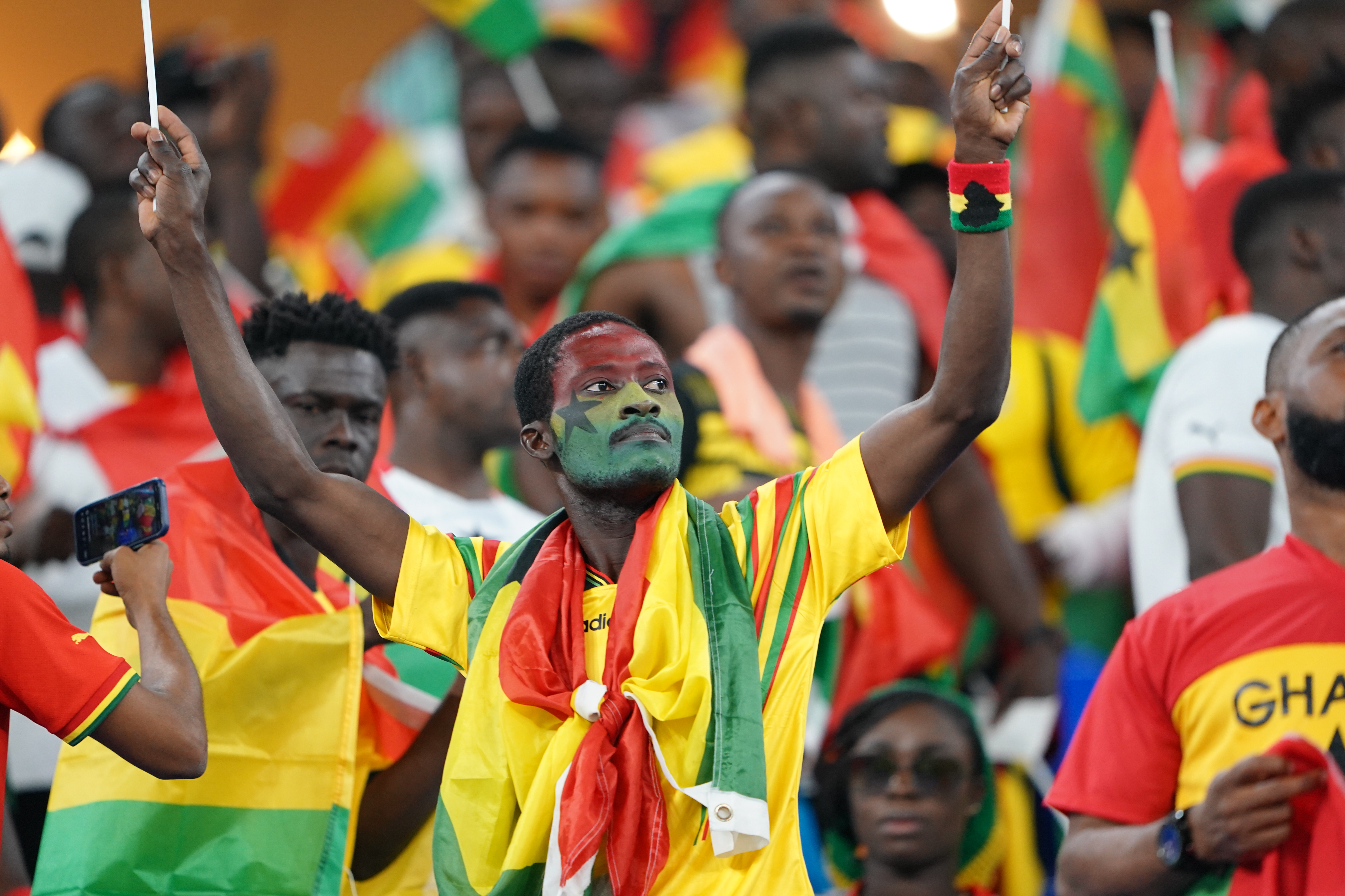 Fan in yellow and green and red clothing and face painted in the same colours holds up flags in each hand with other fans all around him