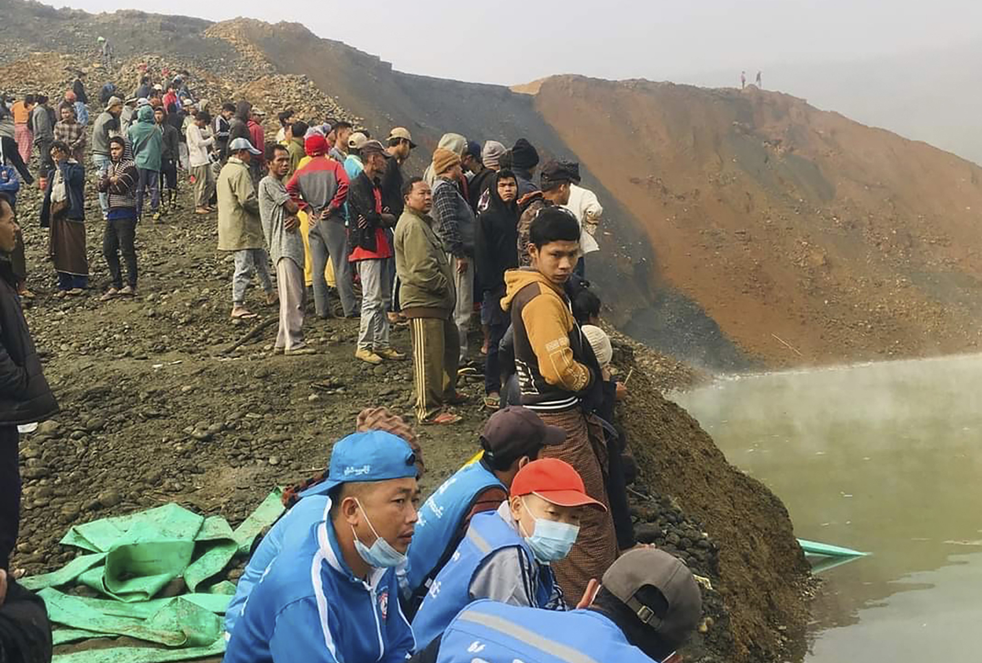 People standing around the rim of a lake formed by gold mining in Hpakant after a landslide. Many look cold and worried.