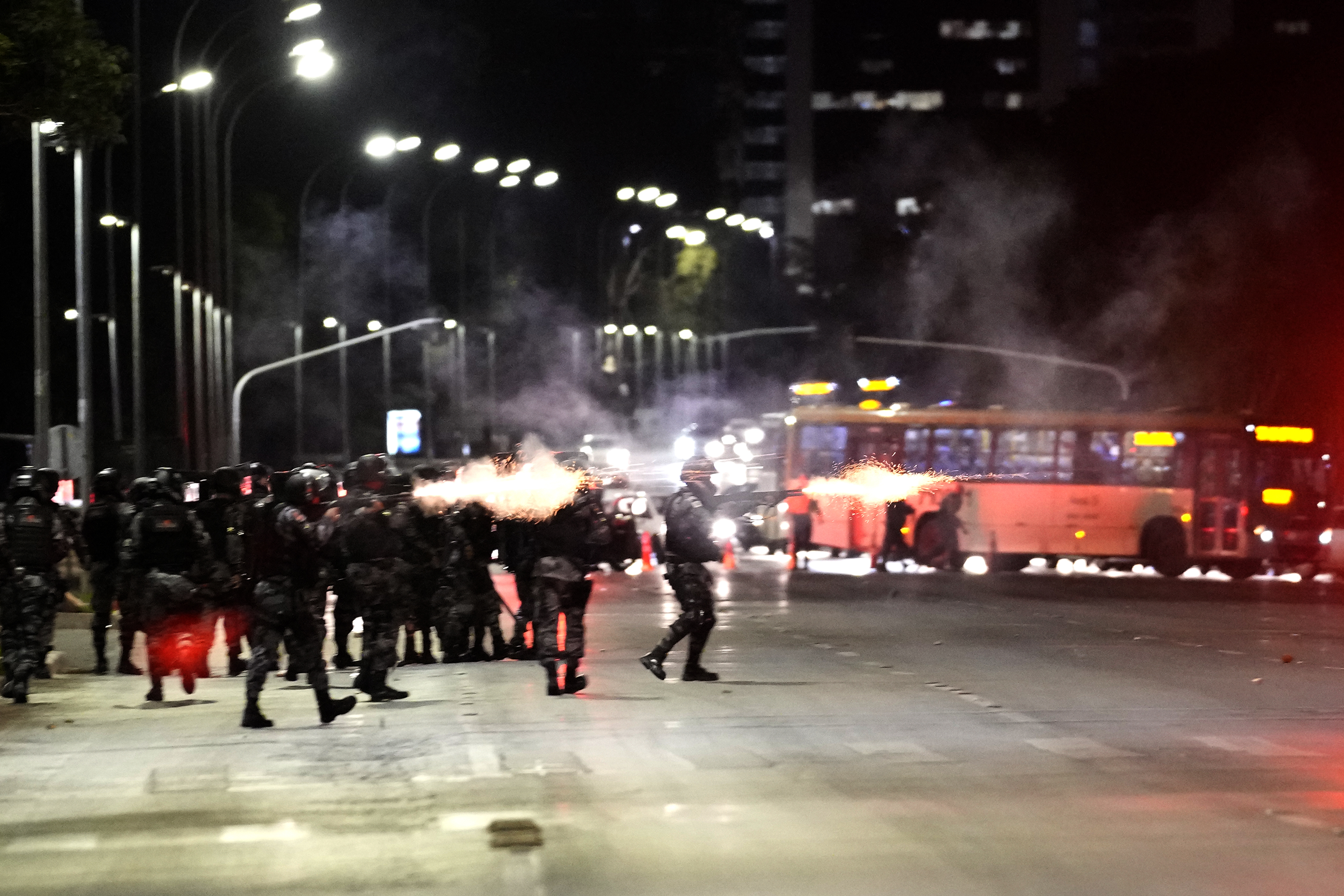 Police and Bolsonaro supporters clash on the street. There is smoke from what are presumably guns being fired to send tear gas canisters in the direction of protesters. The scene looks chaotic.