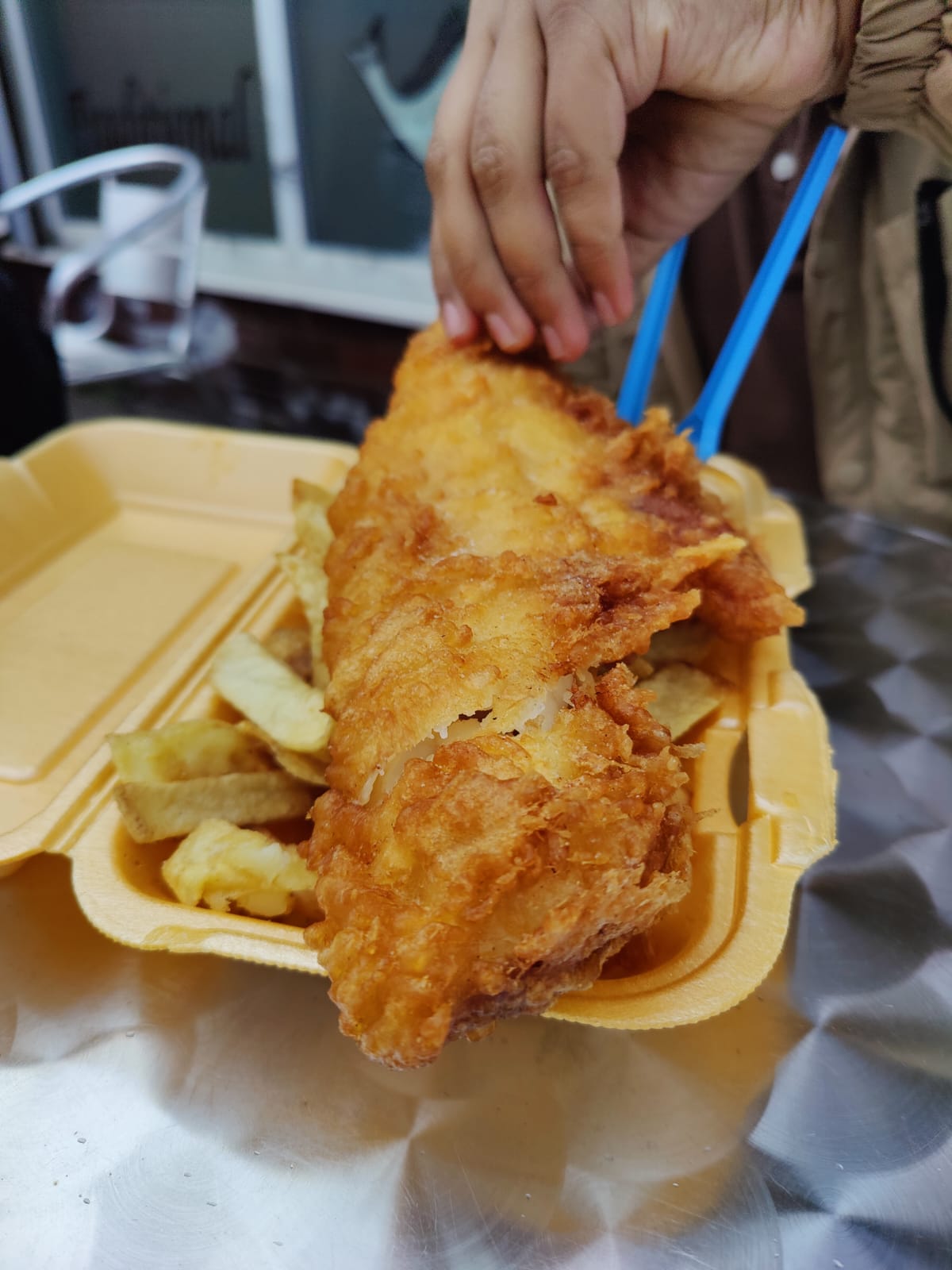 A closeup of a takeout container of fish and chips with Boba's hand grabbing the battered fish to start eating