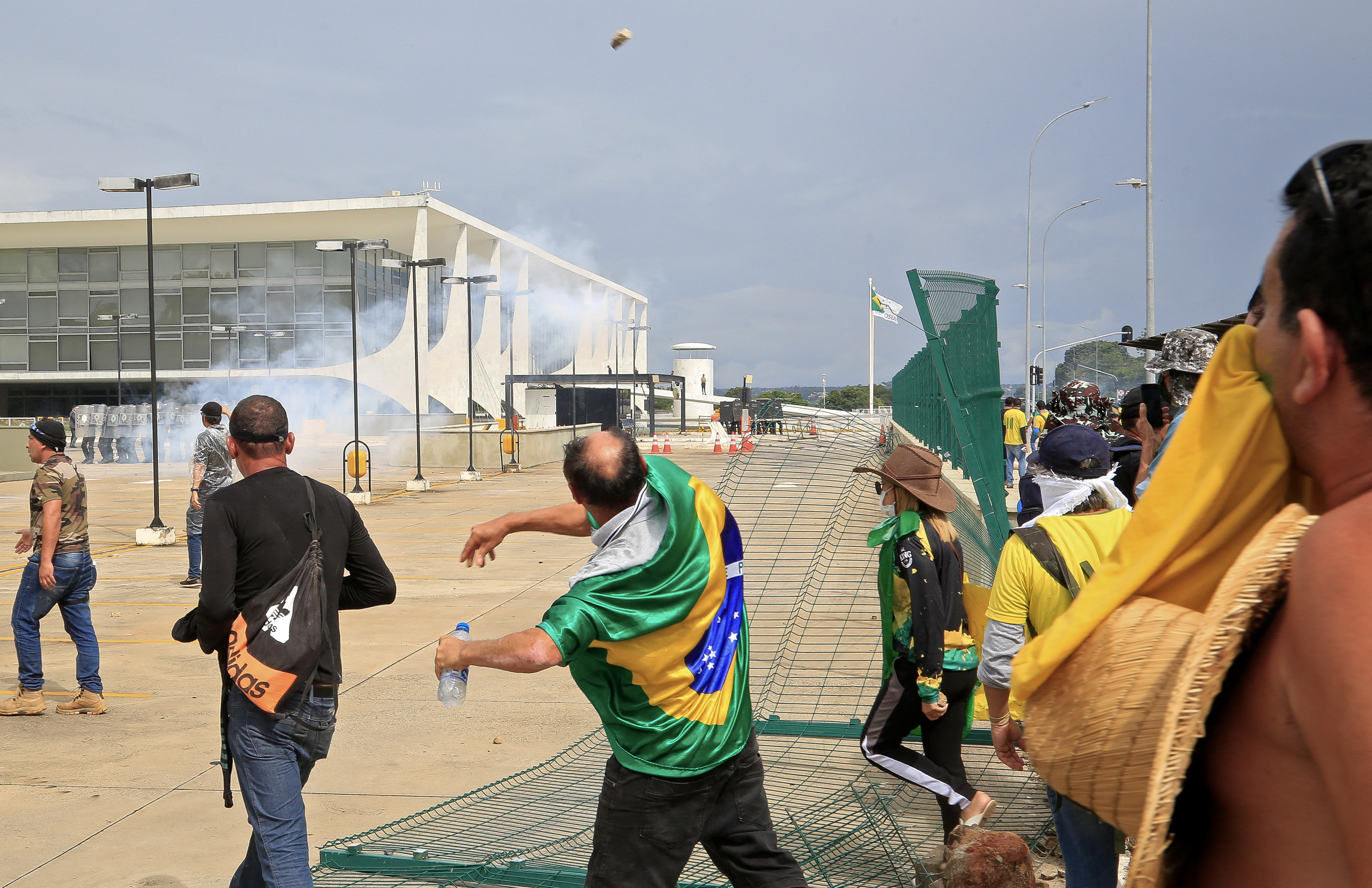 A man with a Brazilian flag around his shoulders throws something towards riot police in Brasilia, Brazil, as Bolsonaro supporters storm key institutions. There is tear gas smoke around them.