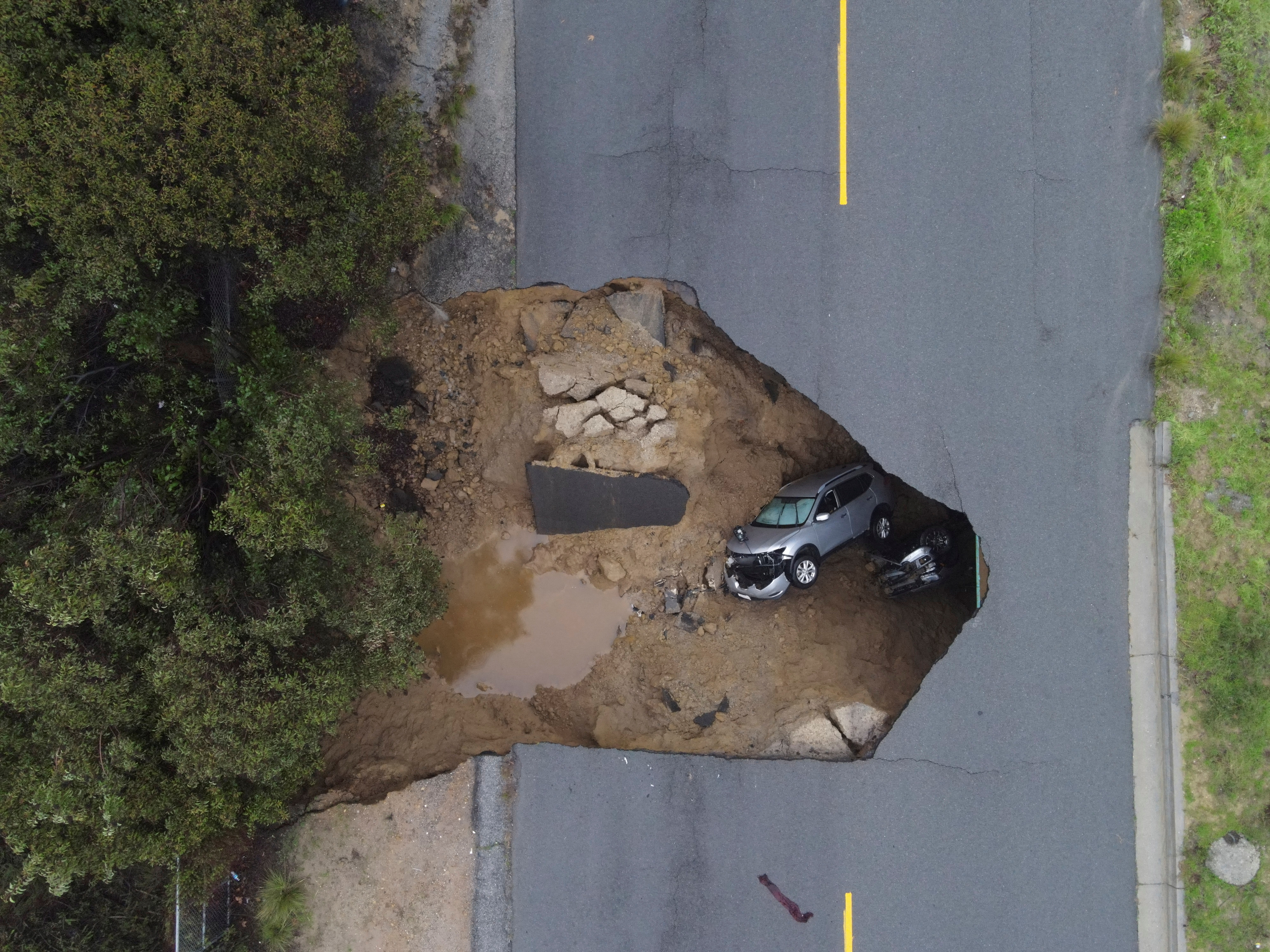 An aerial view of a roadway in California that has partially collapsed, with two vehicles visible under the broken pavement