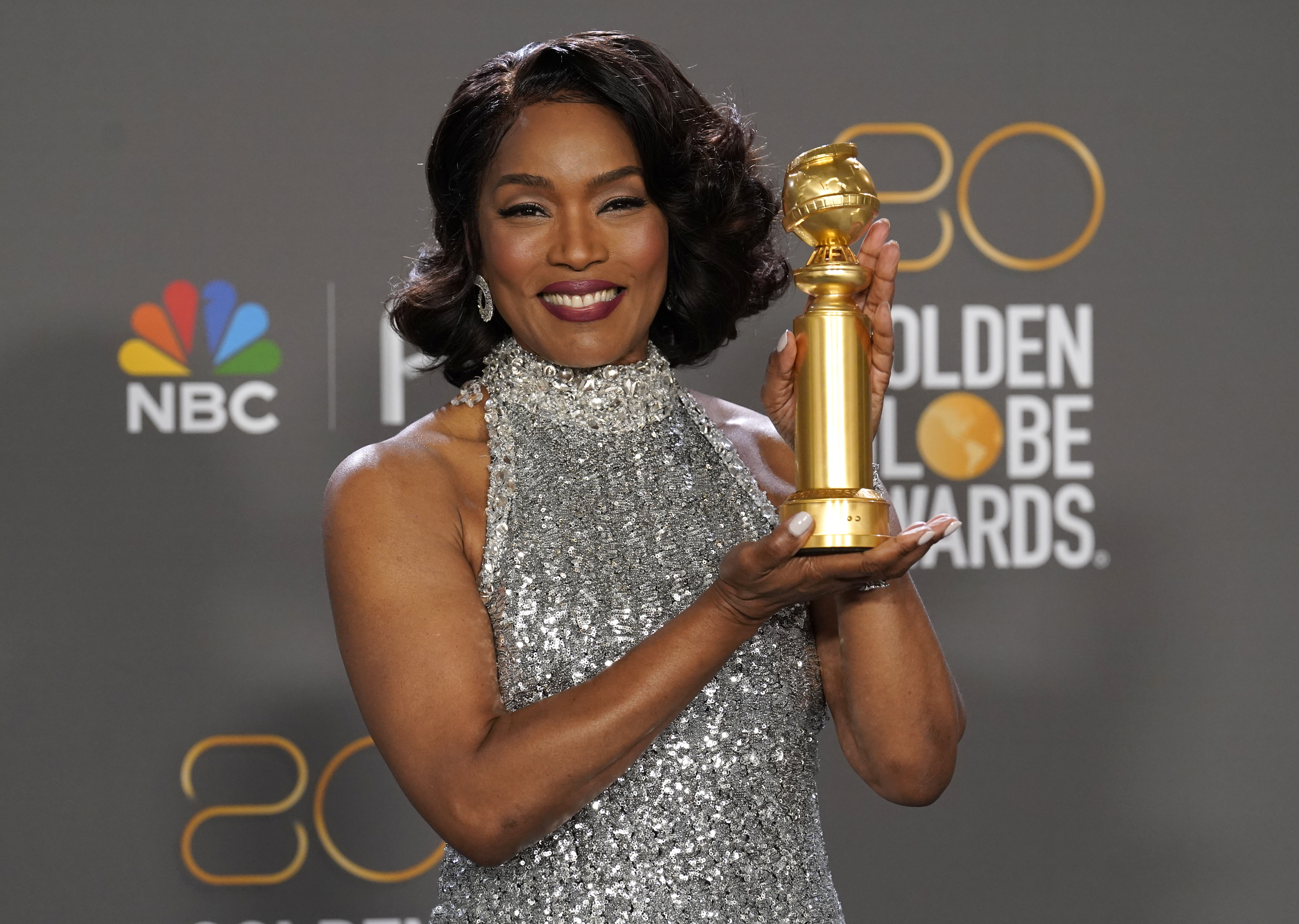 Angela Bassett in a sliver sleeveless high necked evening gown holding her Golden Globe with both hands and smiling.