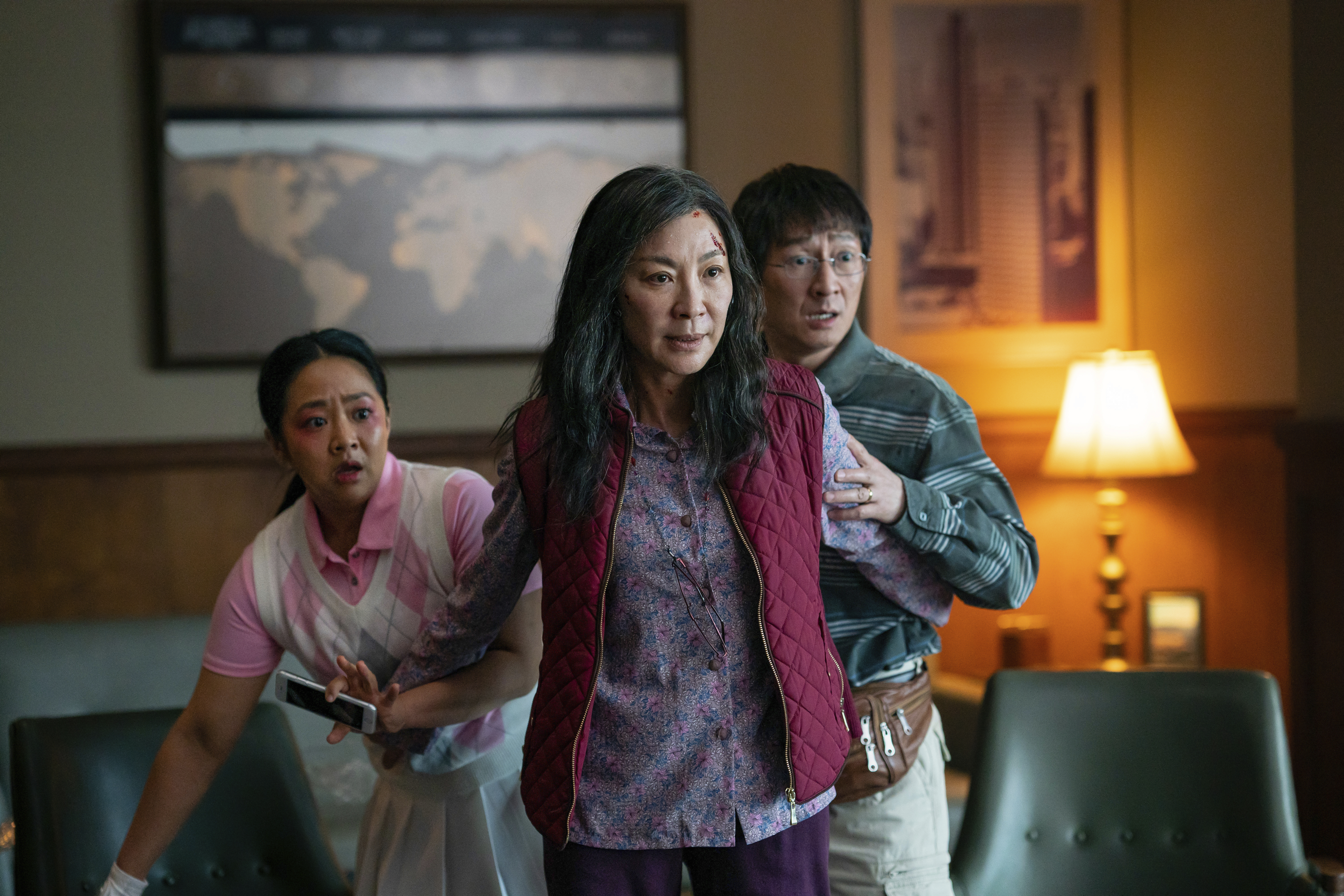A still from Everything Everywhere All At Once showing Stephanie Hsu, Michelle Yeoh and Ke Huy Quan. They are in a living room. Yeoh's character appears to be protecting the others who are standing behind her looking worried