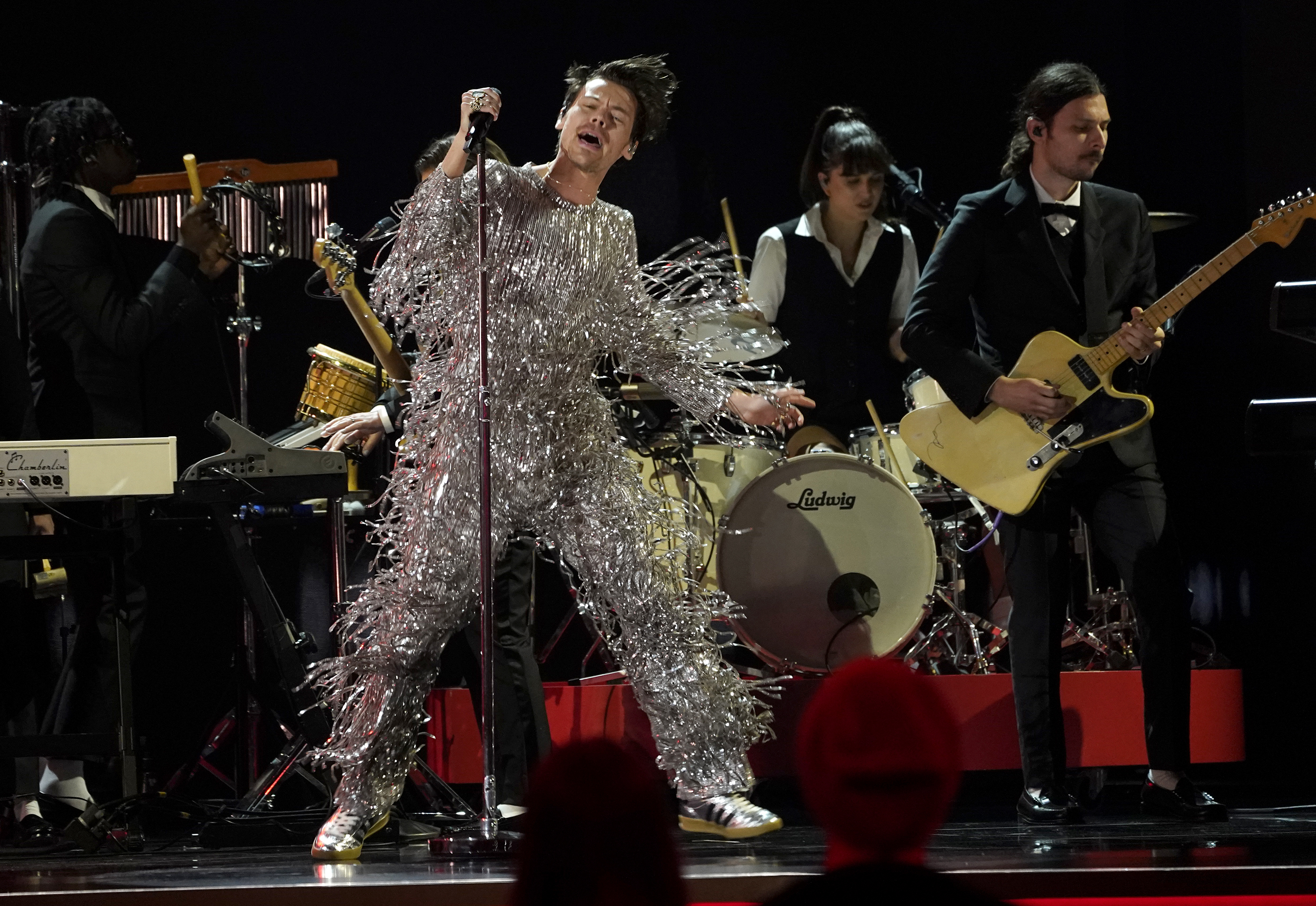 Harry Styles singing As it Was at the Grammys. He is wearing a jumpsuit of silver fringed lame and is dancing, There's a band behind him