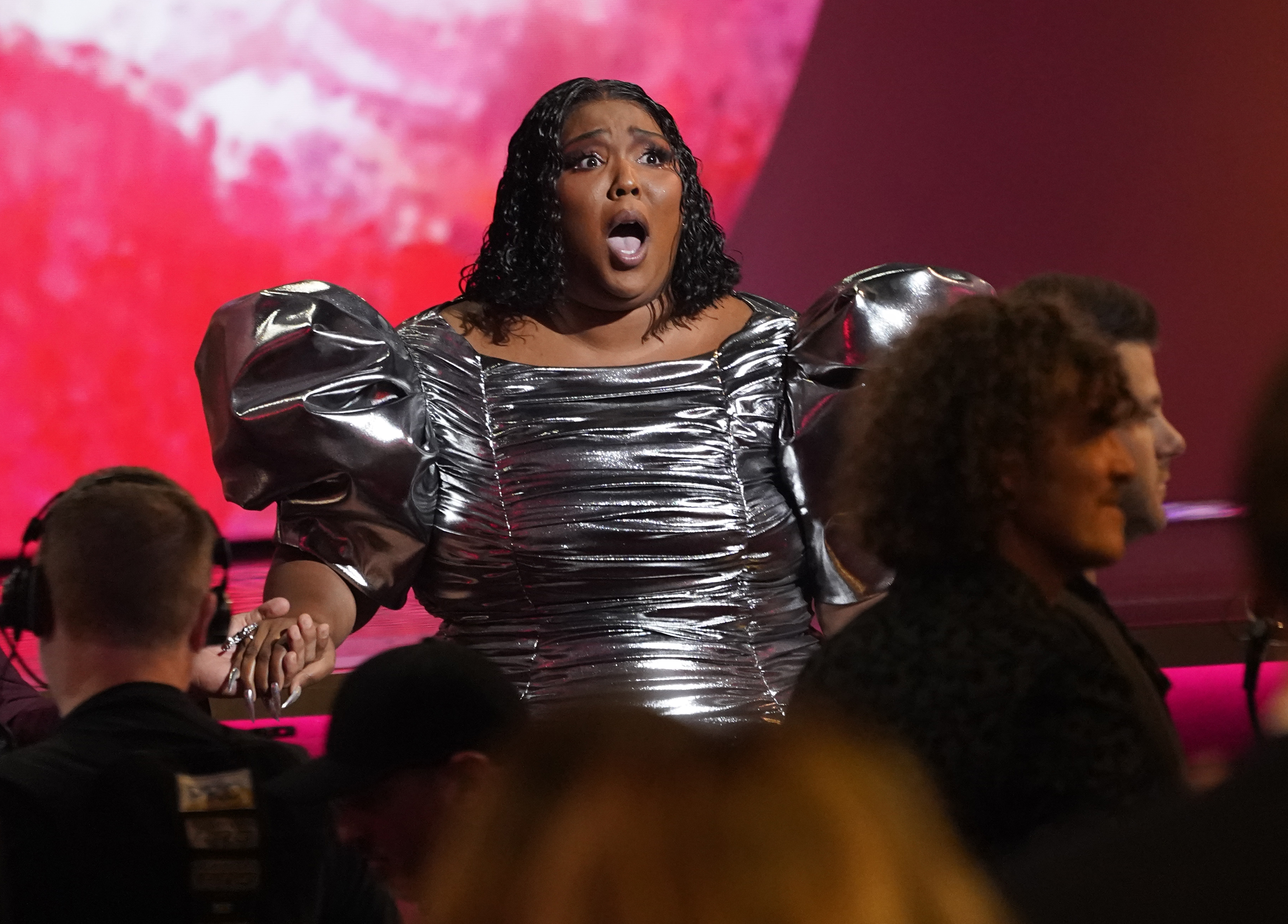 Lizzo is awarded her Grammy for Record of the Year. She is wearing a dark silver dress with big, puffy sleevs and her dark hair is loose. She looks shocked and is gripping someone's hand.