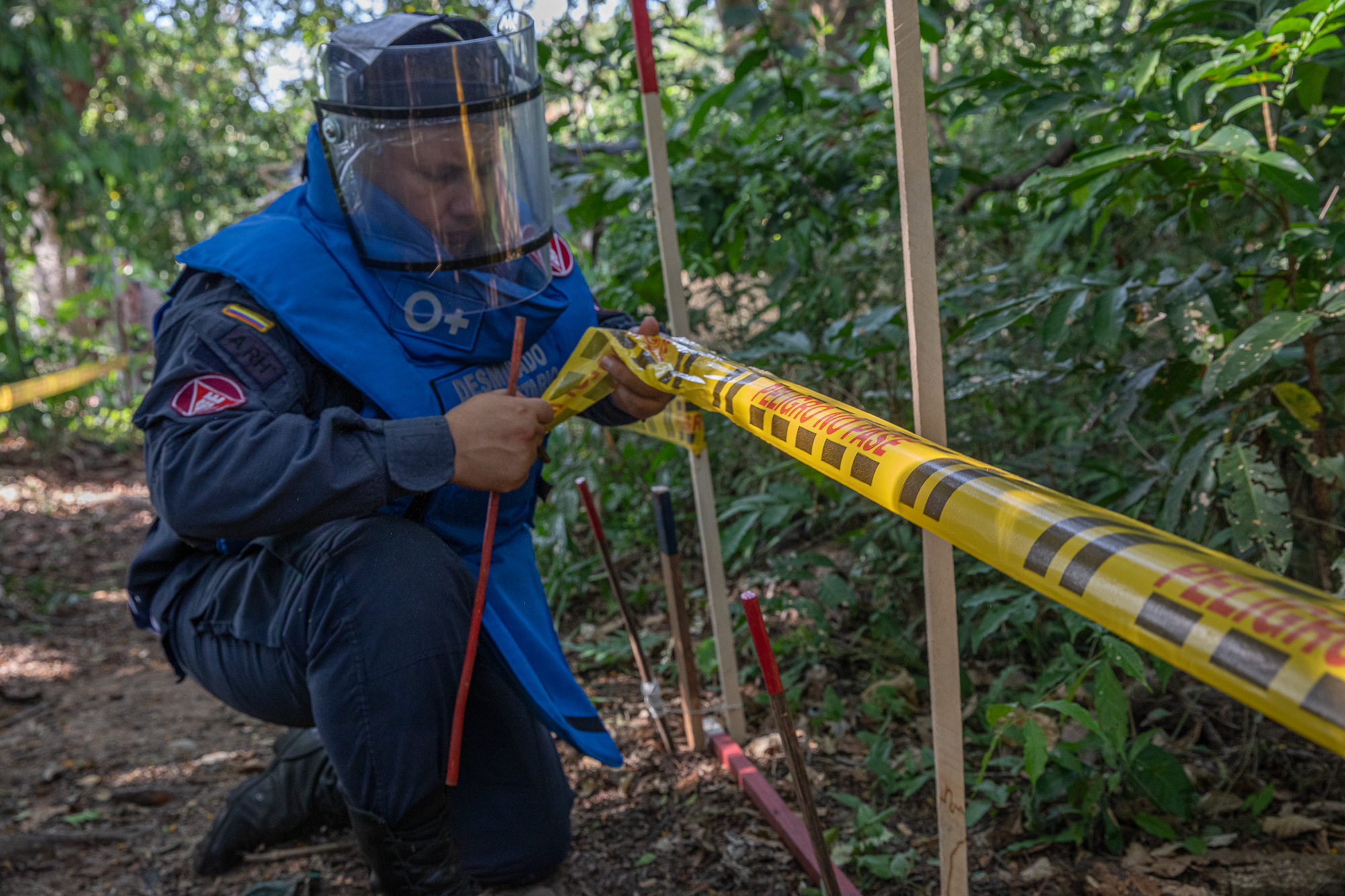 A man wearing blast gear — including a clear shield to protect his face — sets a perimeter with yellow tape, warning of danger from landmines.