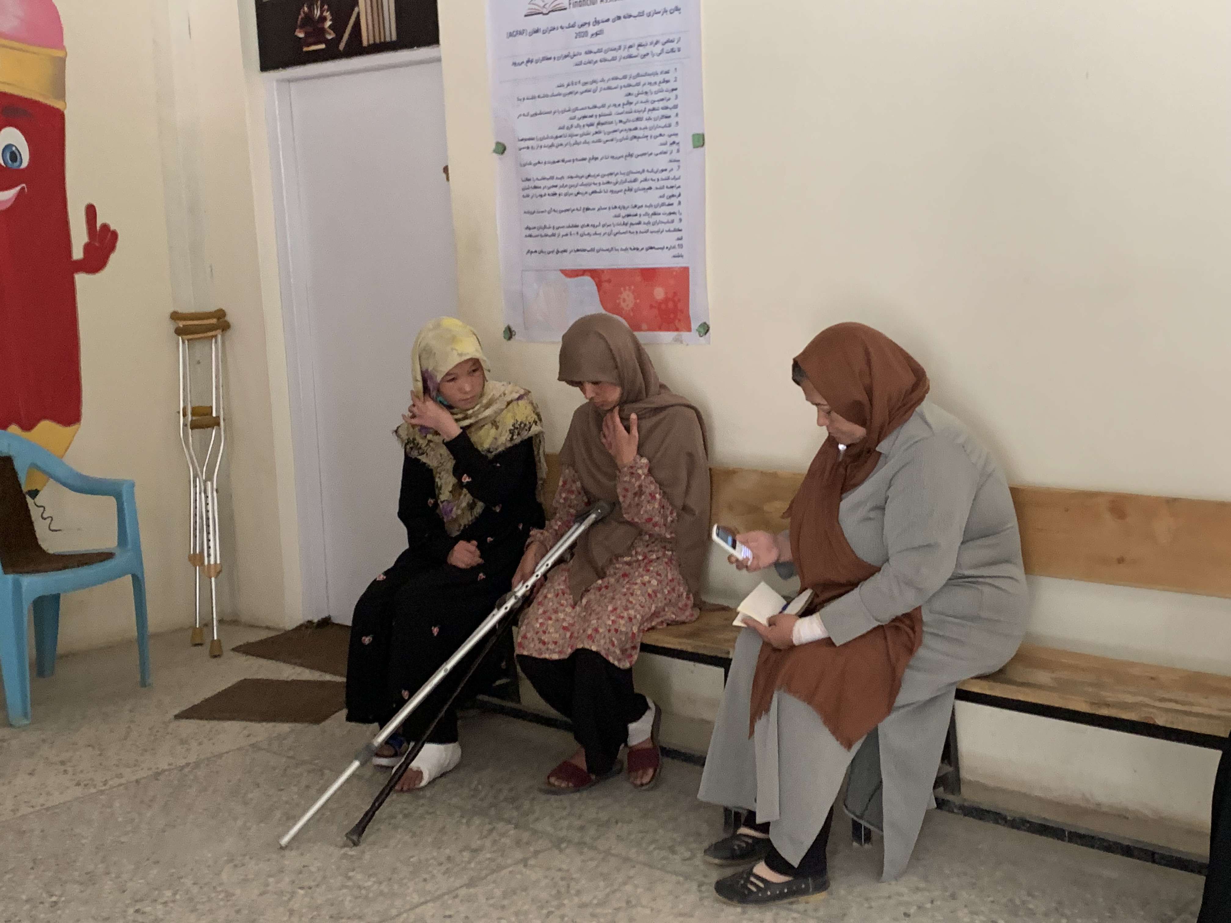 A photo of three women sitting on a bench. the one in the middle is holding a cane and the one on the left has a cast on her foot.