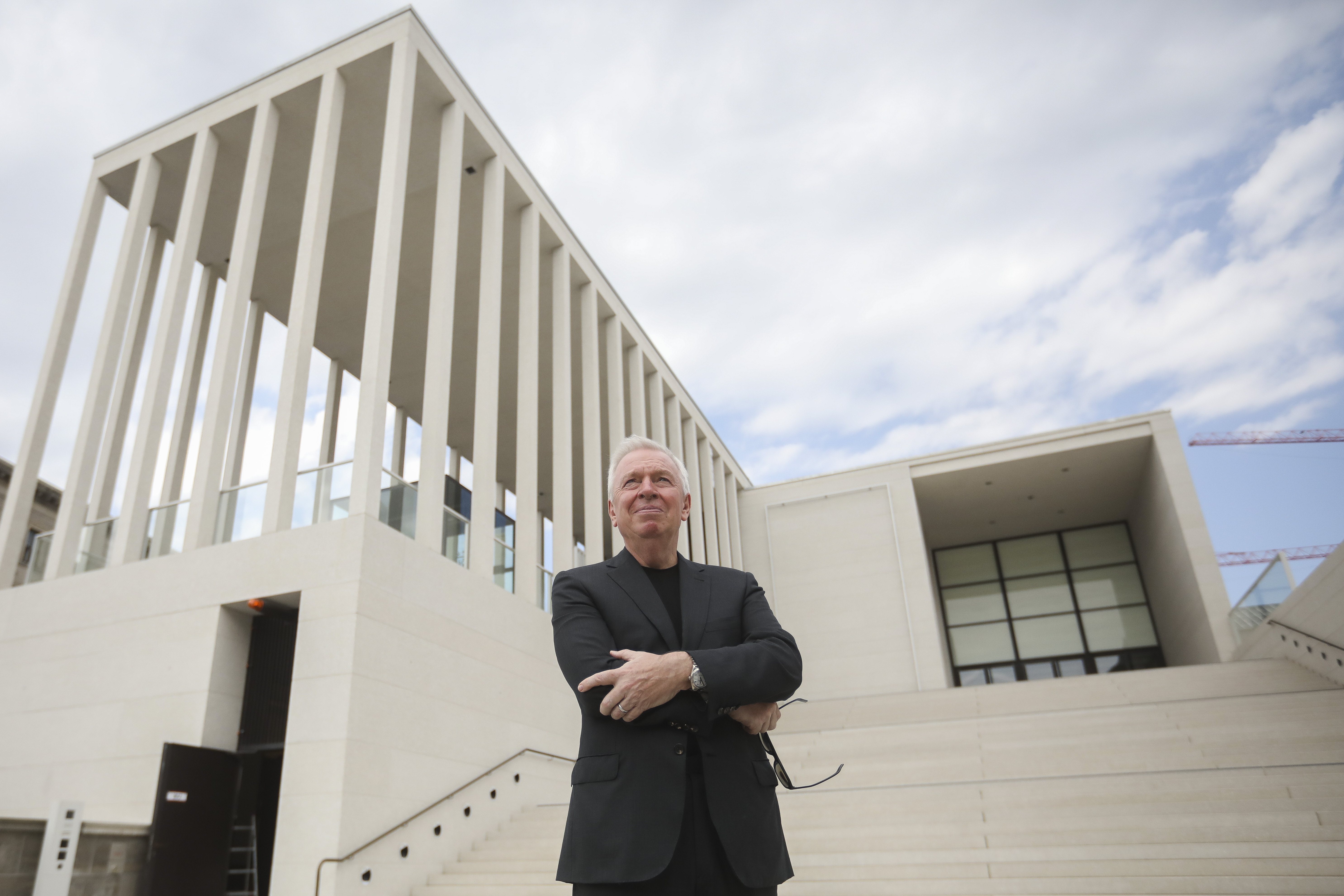 David Chipperfield outside the James-Simon-Galerie at Berlin's Museums Island in Germany. He is standing with his arms crossed at the bottom of some white steps. The building is in an elevated position behind him. It is white and flat-roofed with slender white columns.