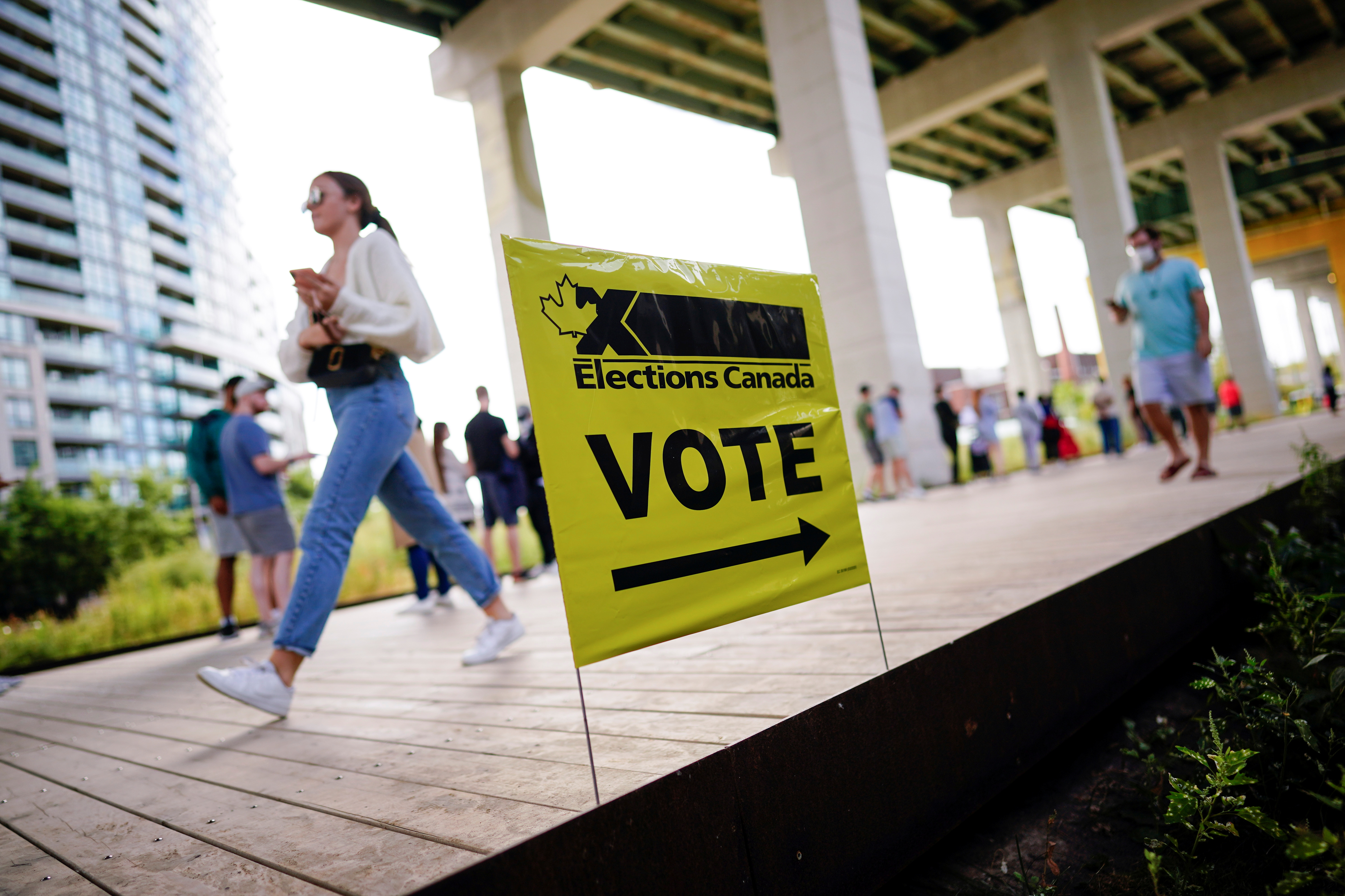 A yellow sign pointing the way to a polling station with the words 'VOTE' written on it. There is a queue of people and a woman striding away in the opposite direction.