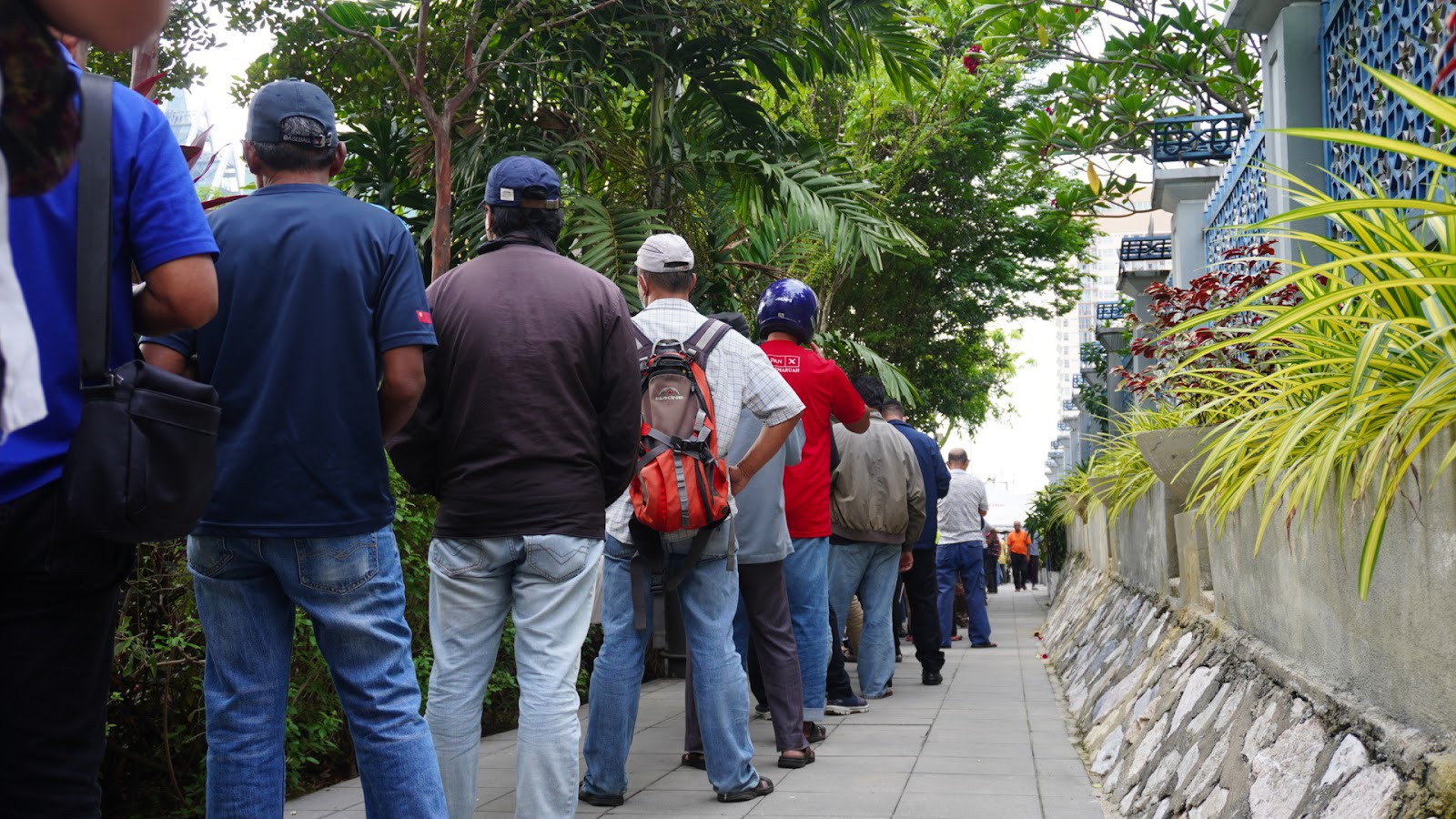 A queue of people outside the mosque waiting for the bubur. The line is moving away from the camera. There is foliage on the right
