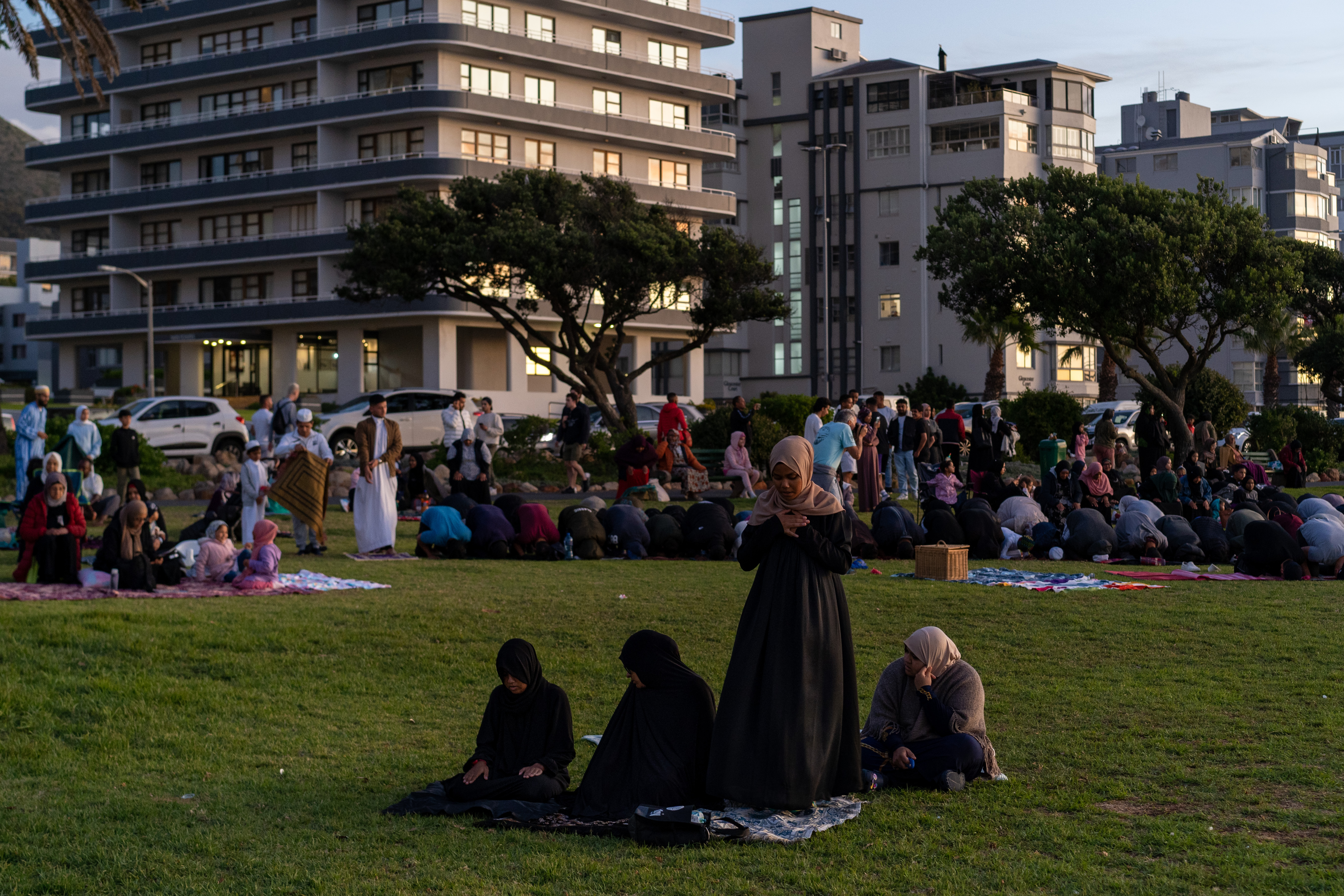 A photo of people sitting on a patch of green with someone praying in the middle.