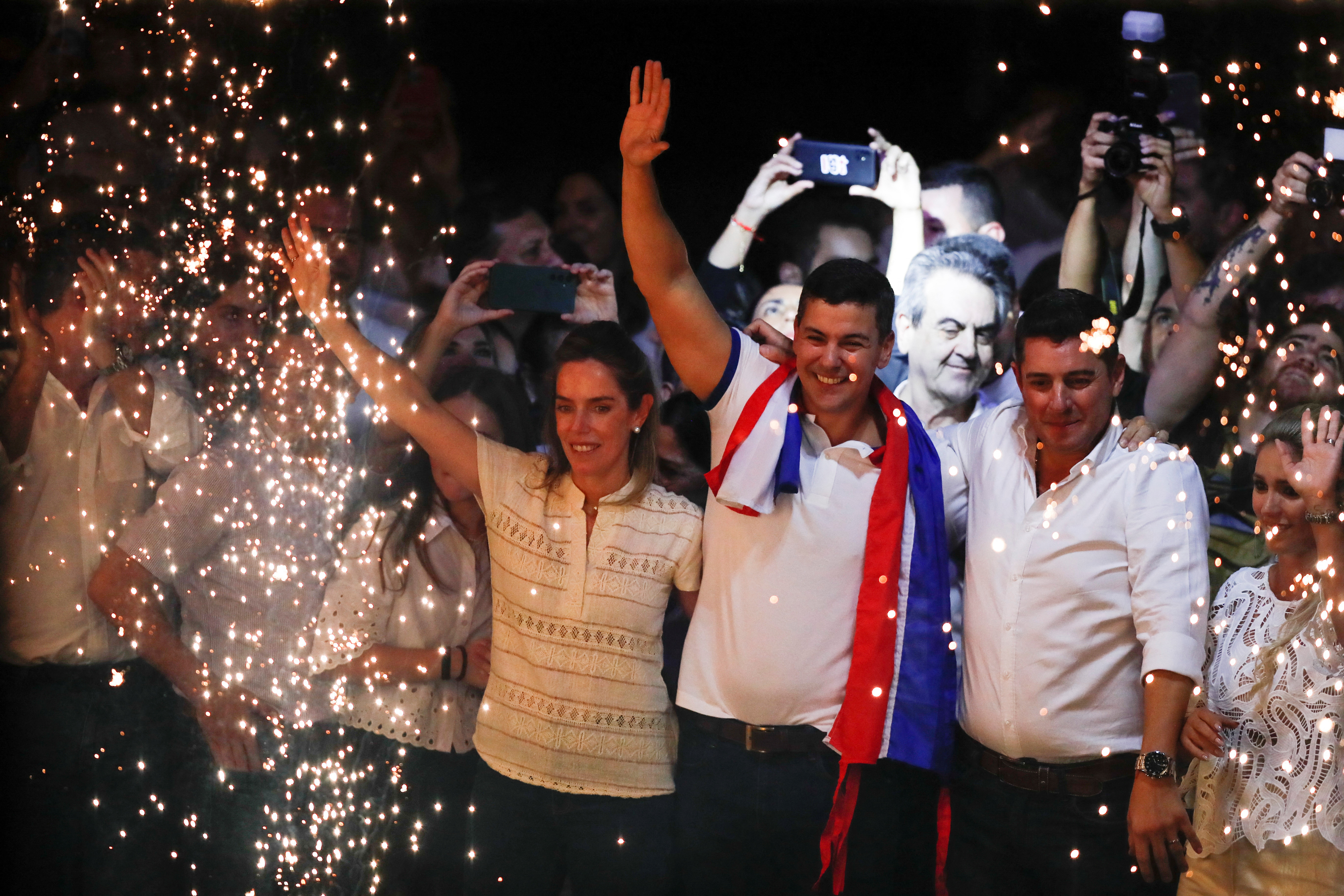 Paraguayan presidential candidate Santiago Pena celebrates his victory. He is standing with his wife, and running mate. They are punching the air. They are surrounded by sparkling light.