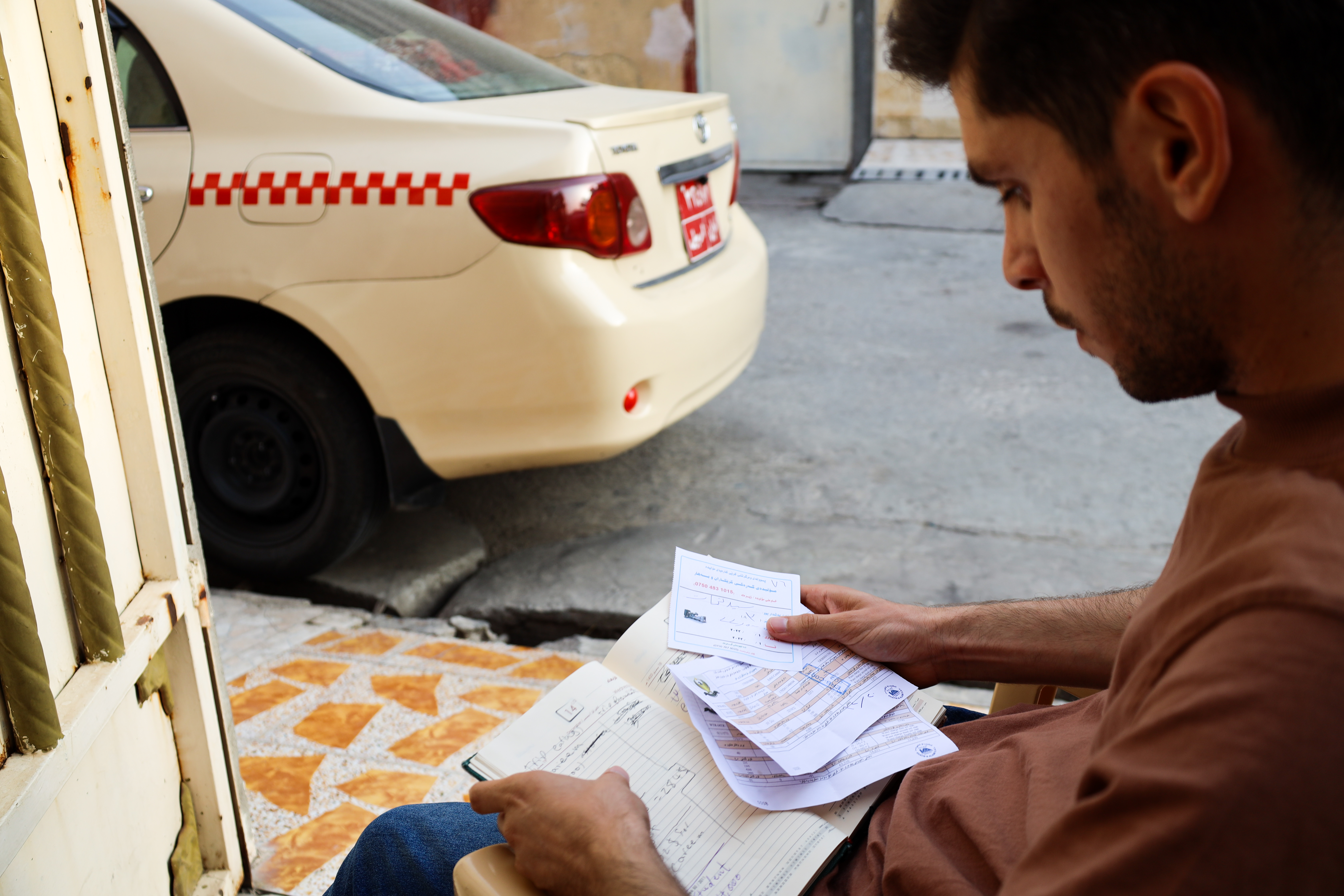 A photo of Mumen looking over his household's April expenses outside with his car in the background.