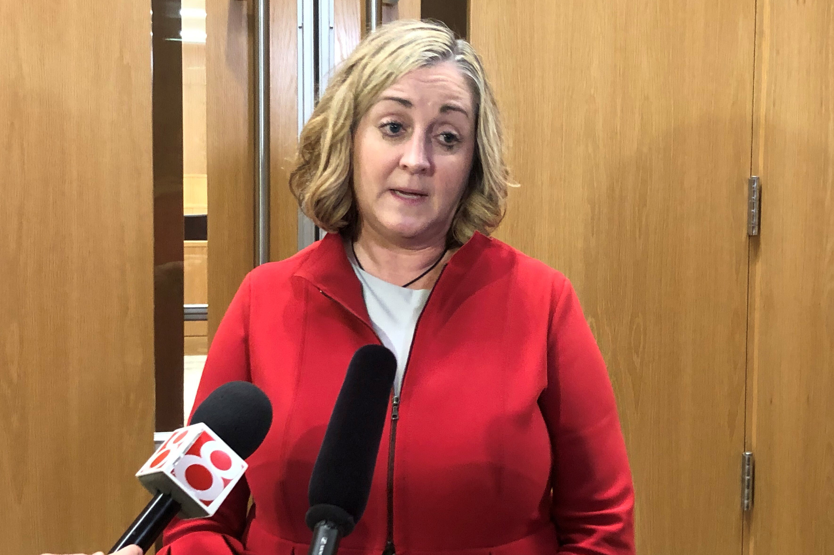 A blonde woman in a red coat speaks into news microphones against a wooden background.