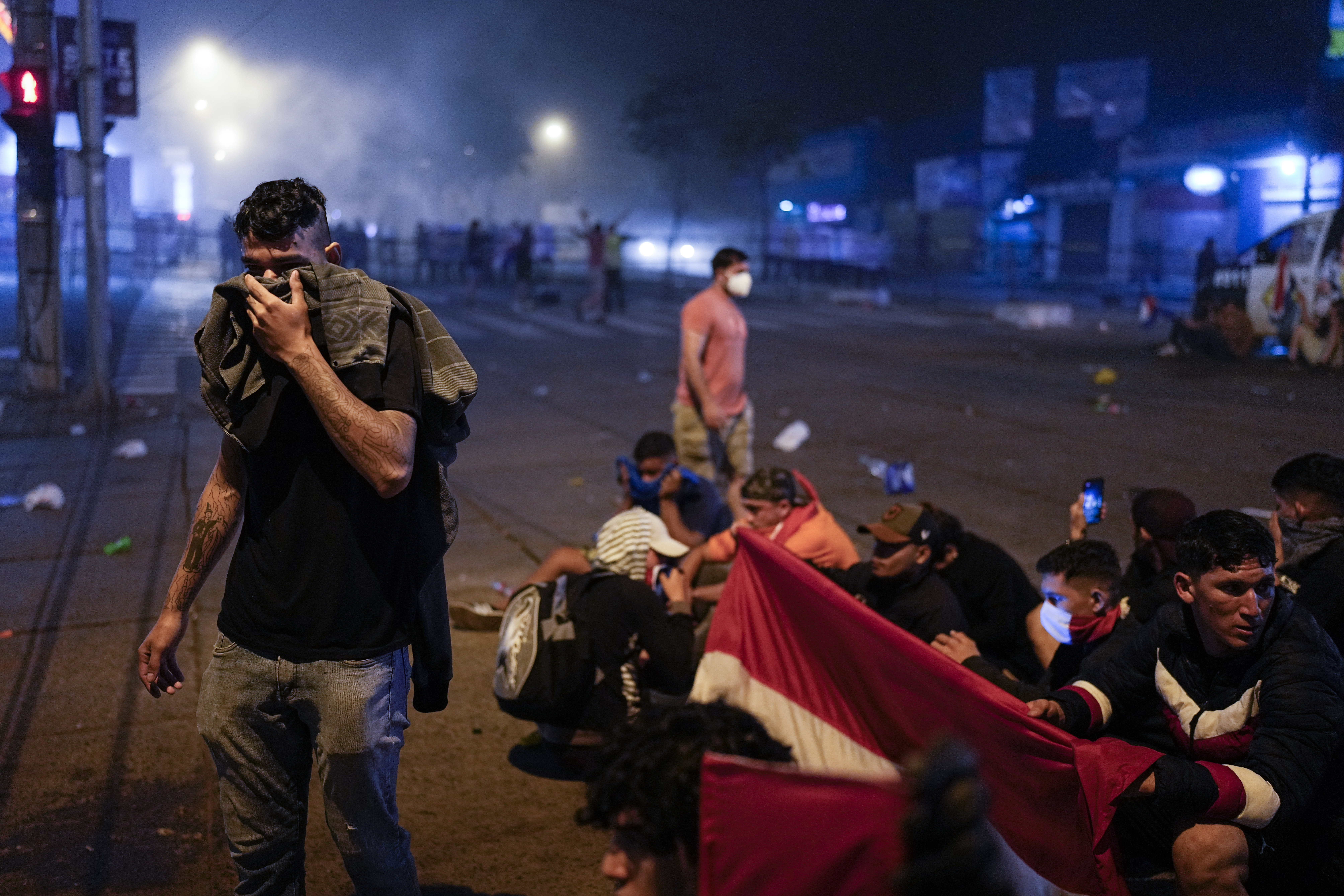 A man covers his face with fabric, while other protesters nearby hold a flag and sit on the ground