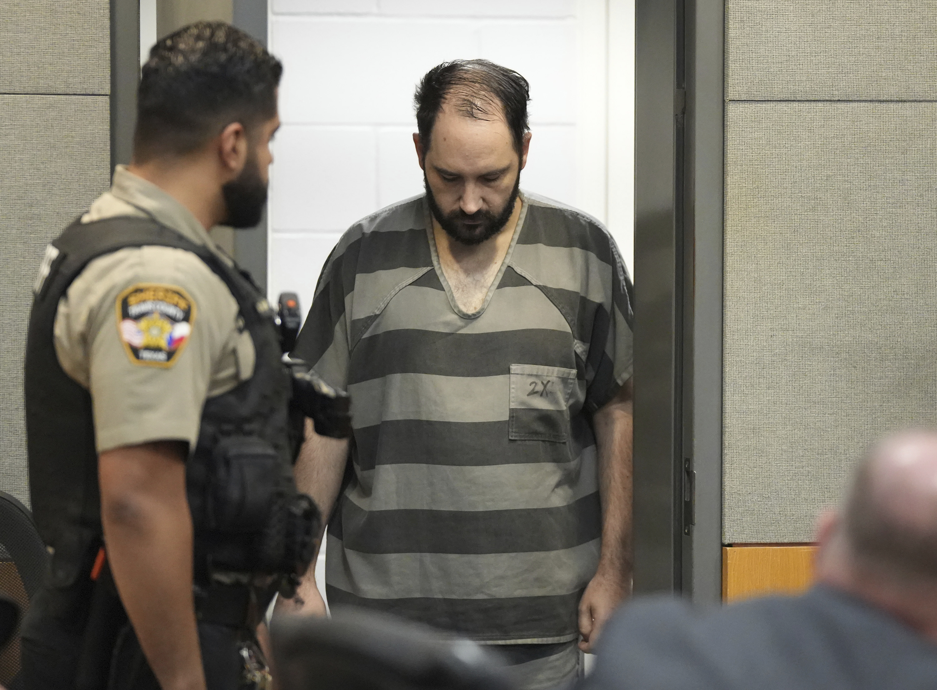 A man in a grey-and-black striped jumpsuit enters a court room, escorted by guards