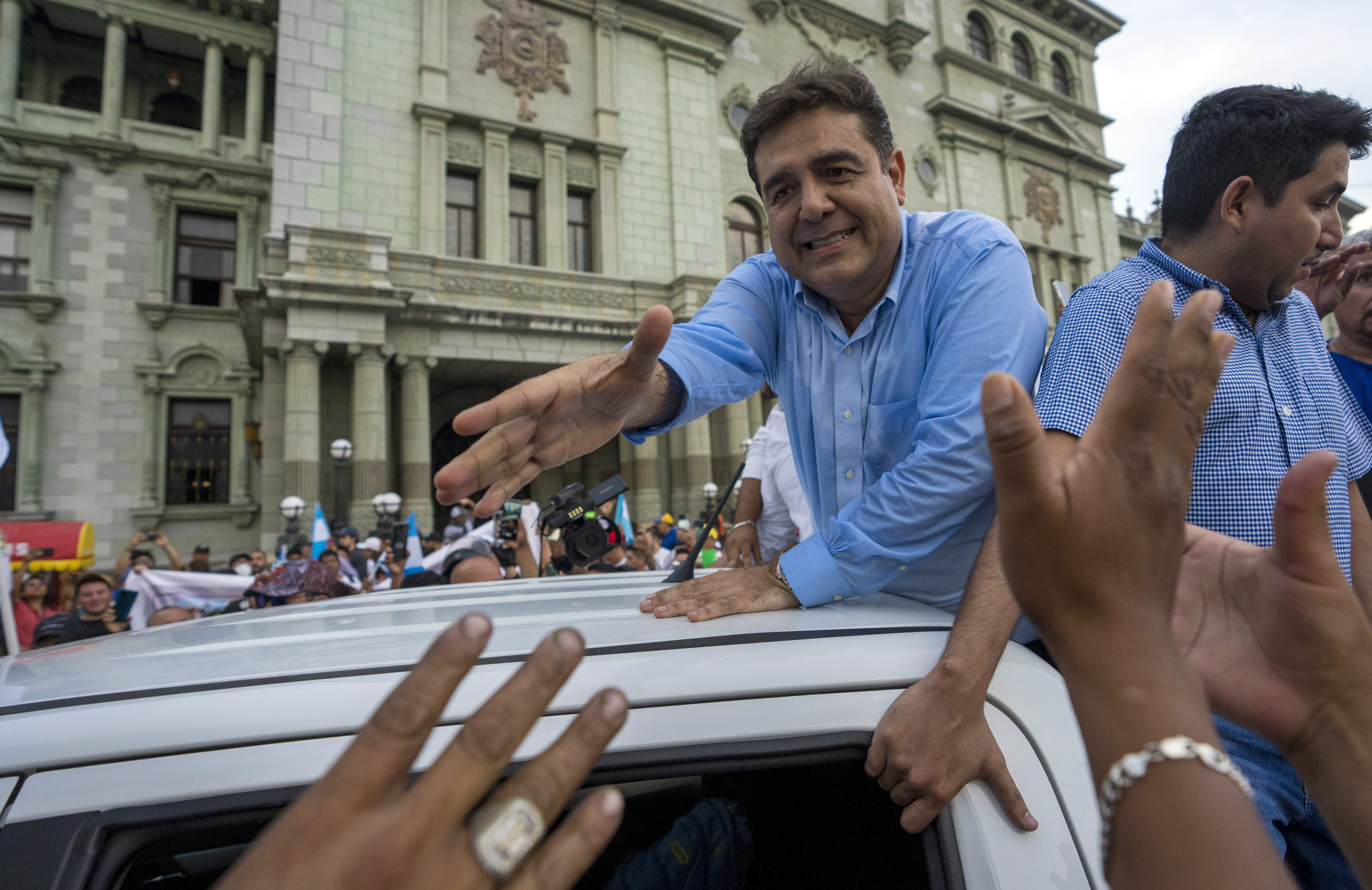 A man, lifting himself above a crowd by propping himself up on a car, reaches out to shake hands with the supporters around him.