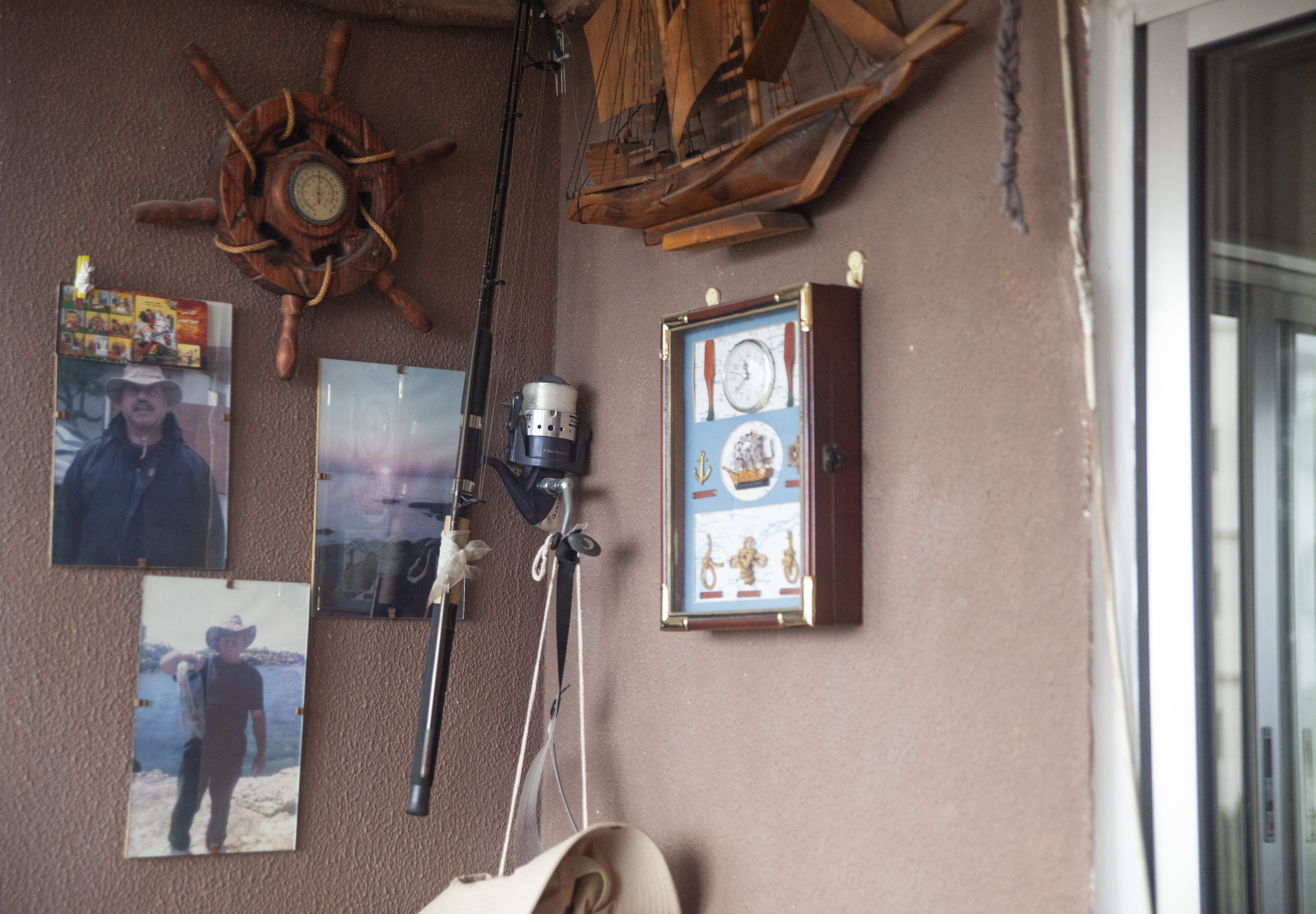 A photo of a corner of a wall with three photos on one end, a ship wheel and a fishing hook and on the other side, a fish net on top with a framed container with three types of knots on it, two fishing flies and a clock.
