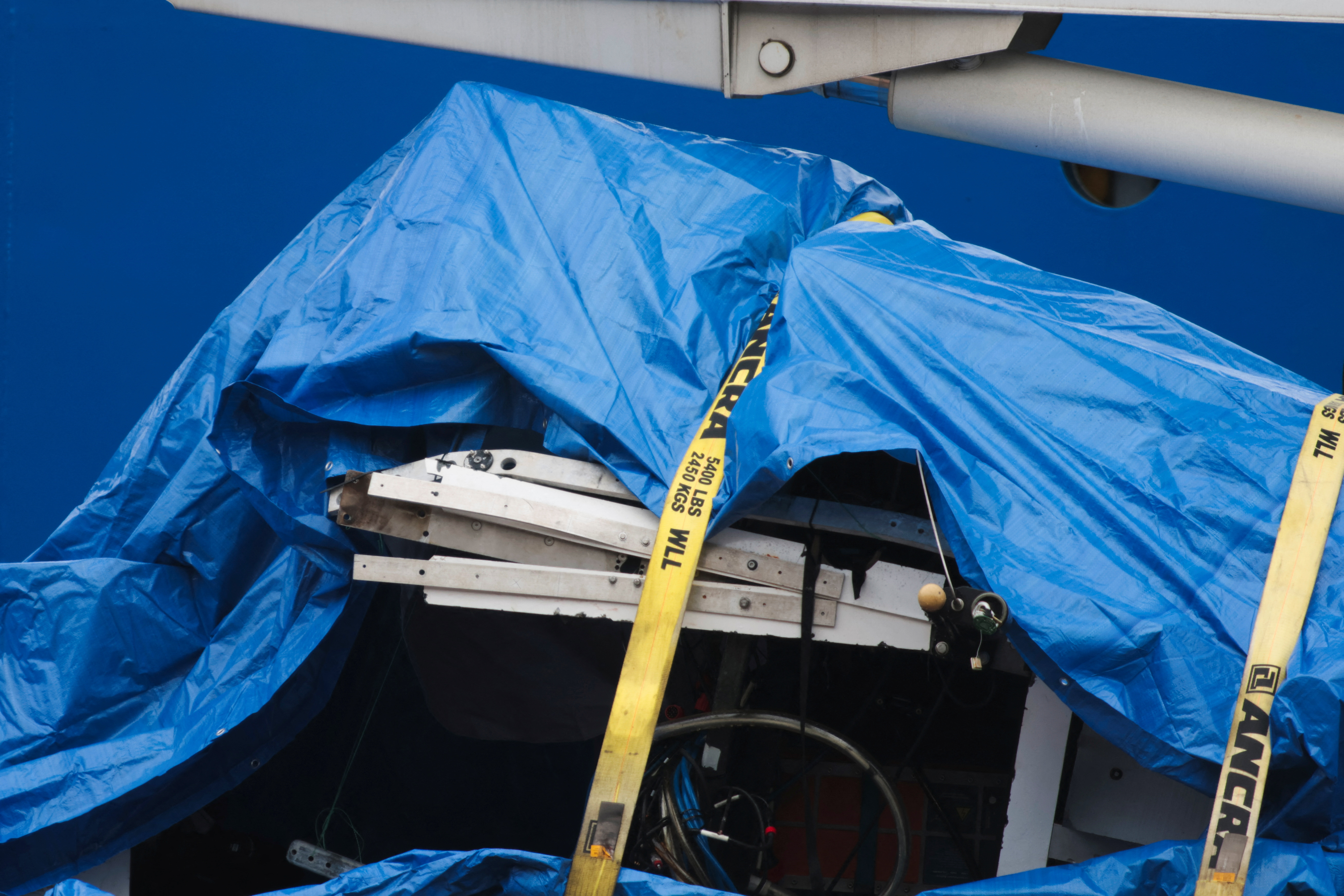 A blue tarp, held down by a yellow strap, covers broken pieces of the sub.